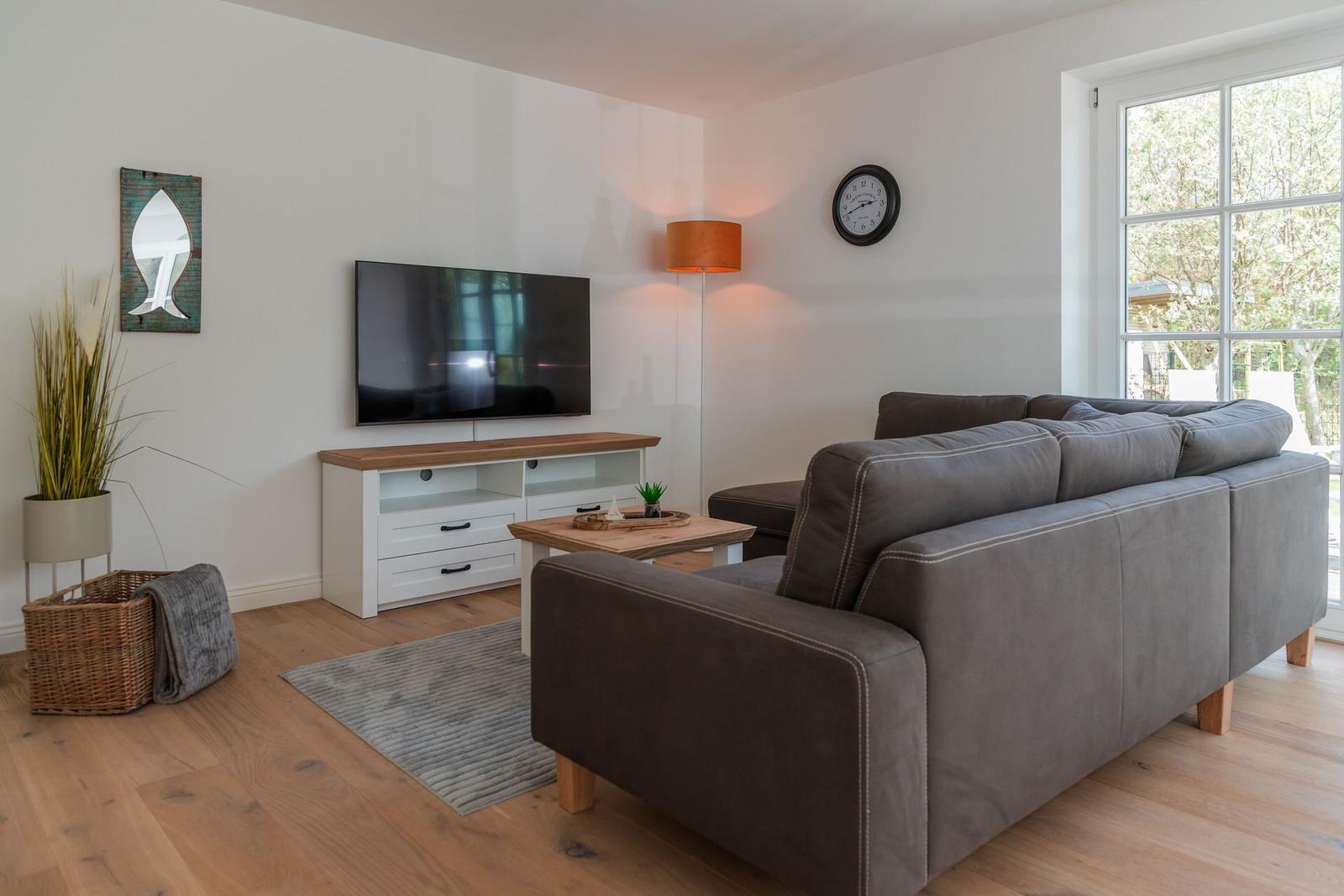 Wohnzimmer mit grauem Sofa, Fernseher und Holzboden. Fenster mit Blick auf Garten.