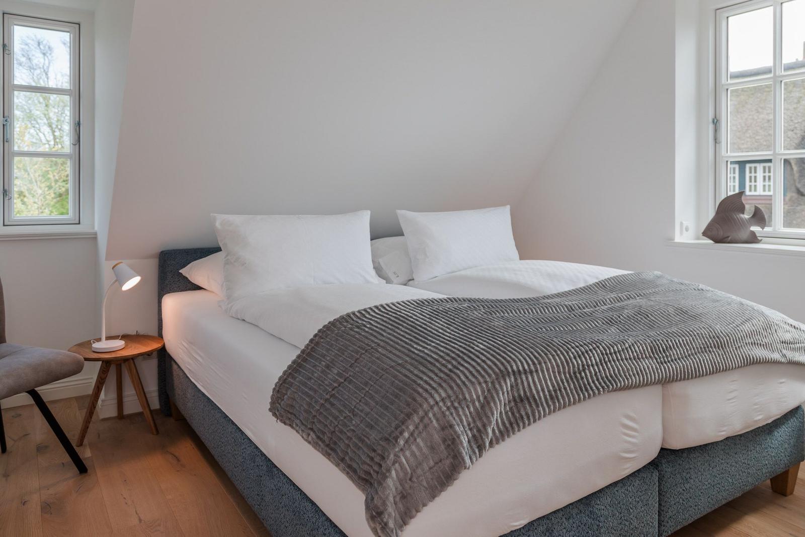 Schlafzimmer mit Doppelbett, zwei Fenstern und Holzboden.