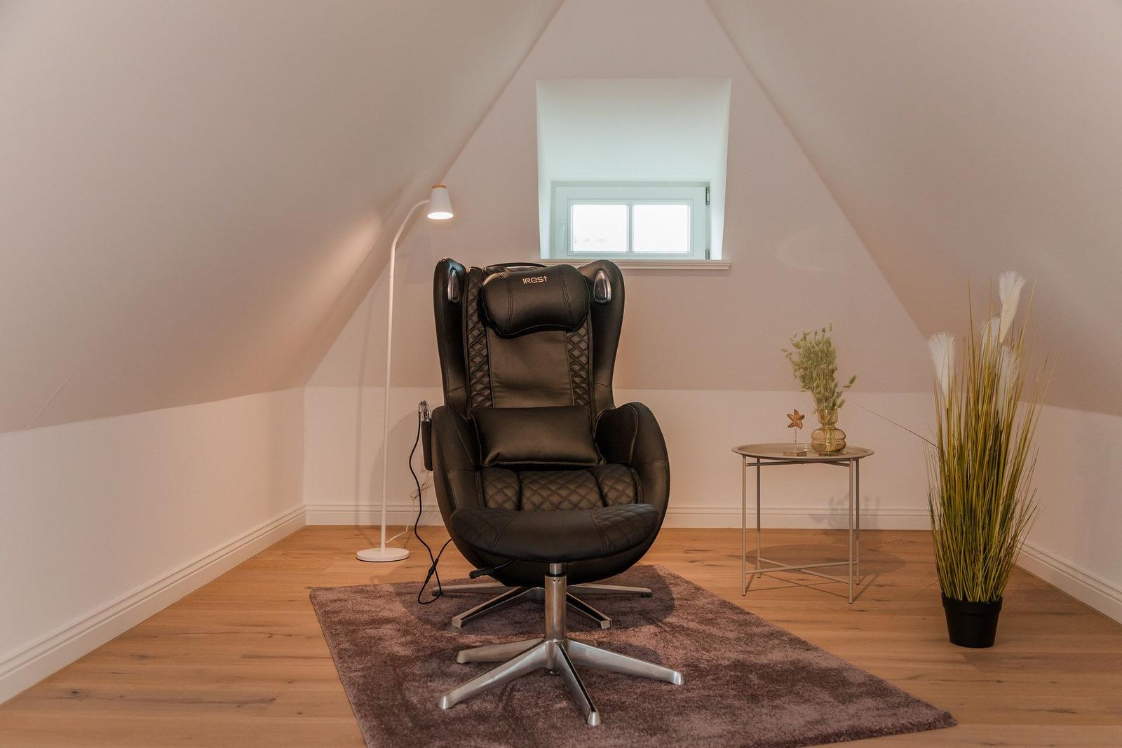 Schwarze Massagestuhl mit Fußstütze in einem Dachzimmer mit Holzboden und Fenster.