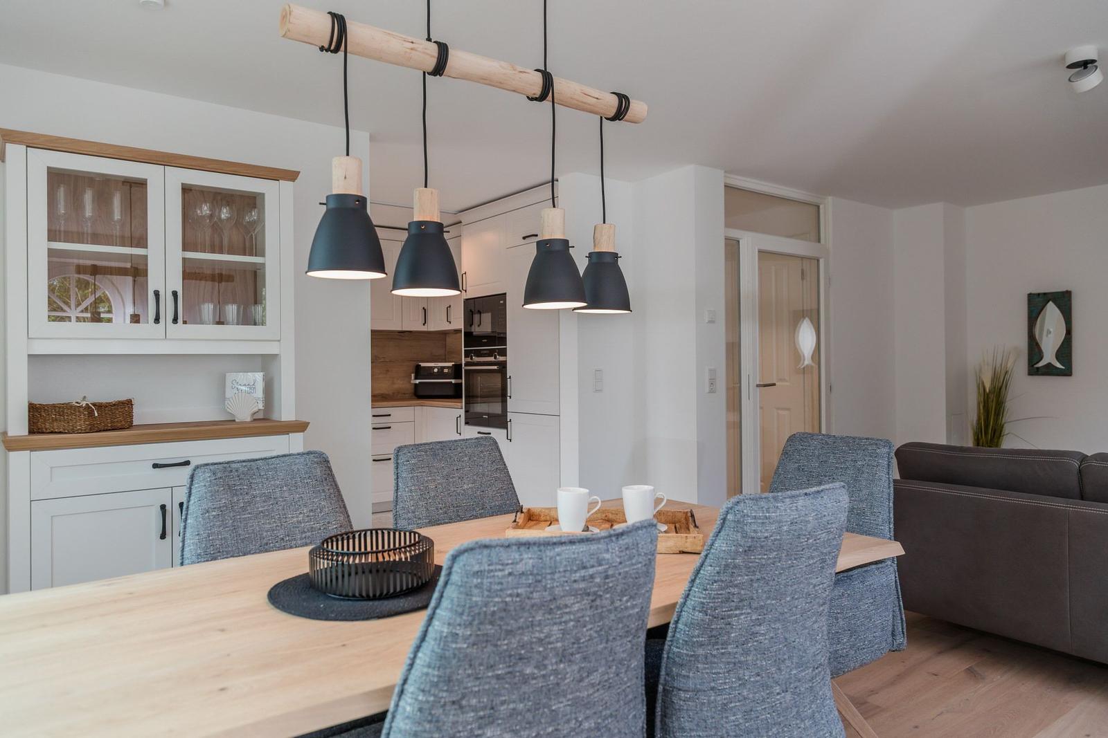 Essbereich mit Holztisch, grauen Stühlen und moderner Deckenleuchte. Küche und Wohnbereich sind sichtbar.