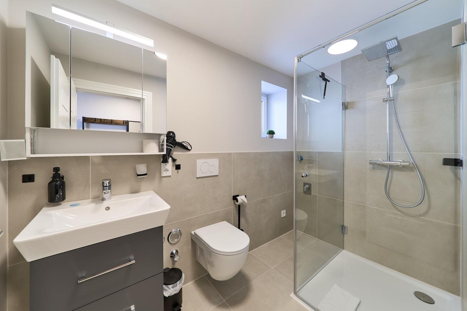 Badezimmer mit Dusche, Waschbecken und Toilette. Moderne Einrichtung mit Spiegel und Fenster.