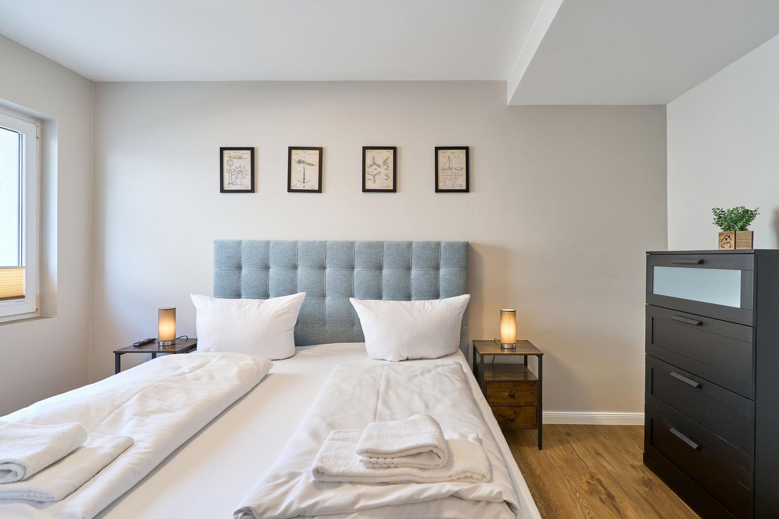 Schlafzimmer mit Bett, Nachttischen und Kleiderschrank. An der Wand hängen vier gerahmte Bilder.