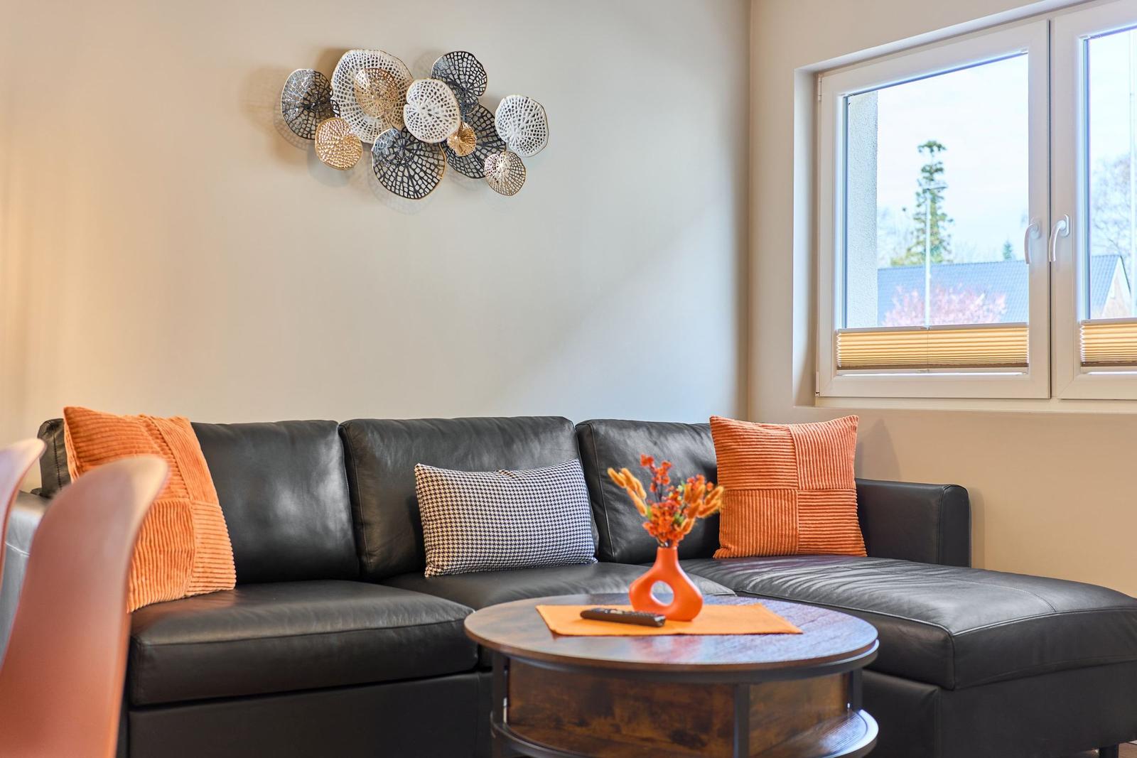 Schwarzes Sofa mit orangefarbenen Kissen und Wanddekoration. Rundes Tischchen mit Blumen.