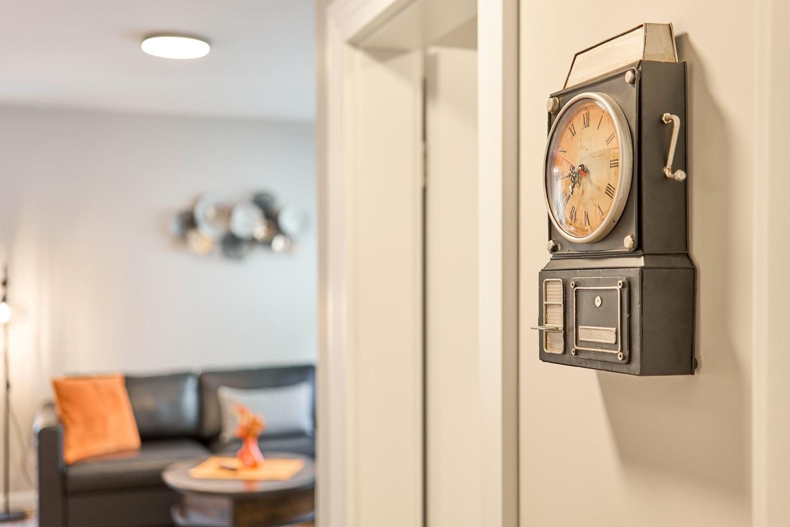 Ein antiker Wanduhr hängt an der Wand. Im Hintergrund ist ein Wohnbereich mit Sofa und Couchtisch.