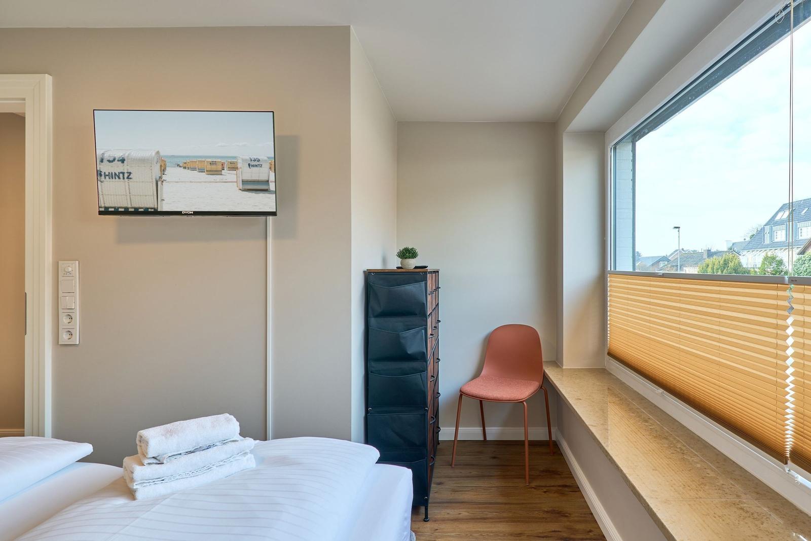 Zimmer mit Bett, Fernseher und Fensterbank. Orange Stuhl und Holzboden.