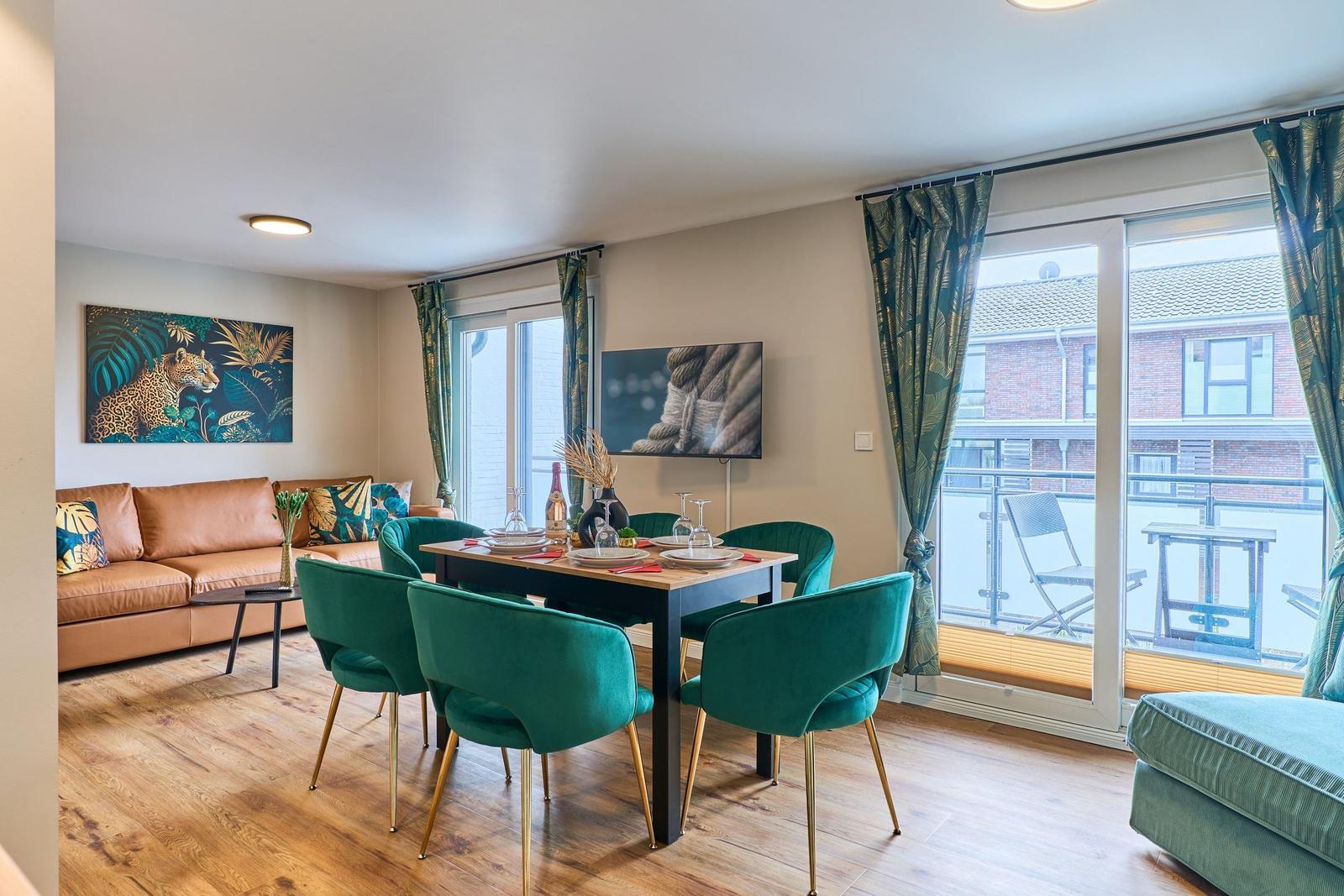 Essbereich mit grünen Stühlen und Holztisch. Wohnbereich mit Sofa und Fernseher. Balkon mit Sitzgelegenheit.