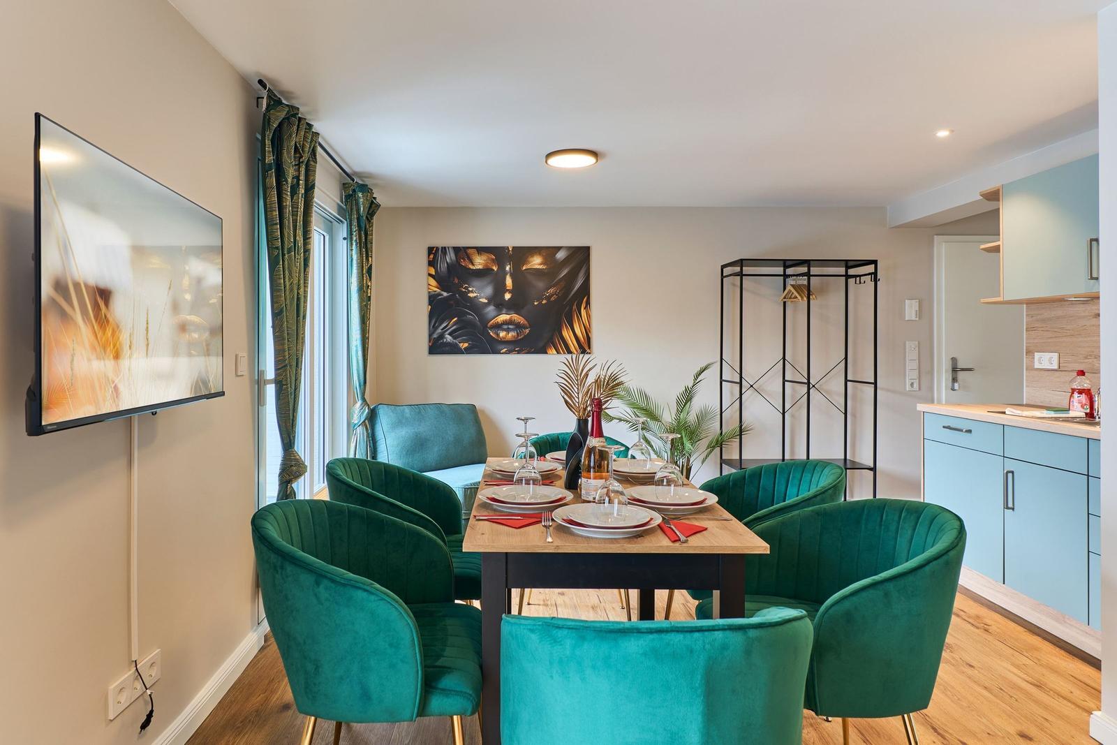 Esszimmer mit grünen Stühlen, Esstisch und Wandbild. Küche und TV sichtbar.