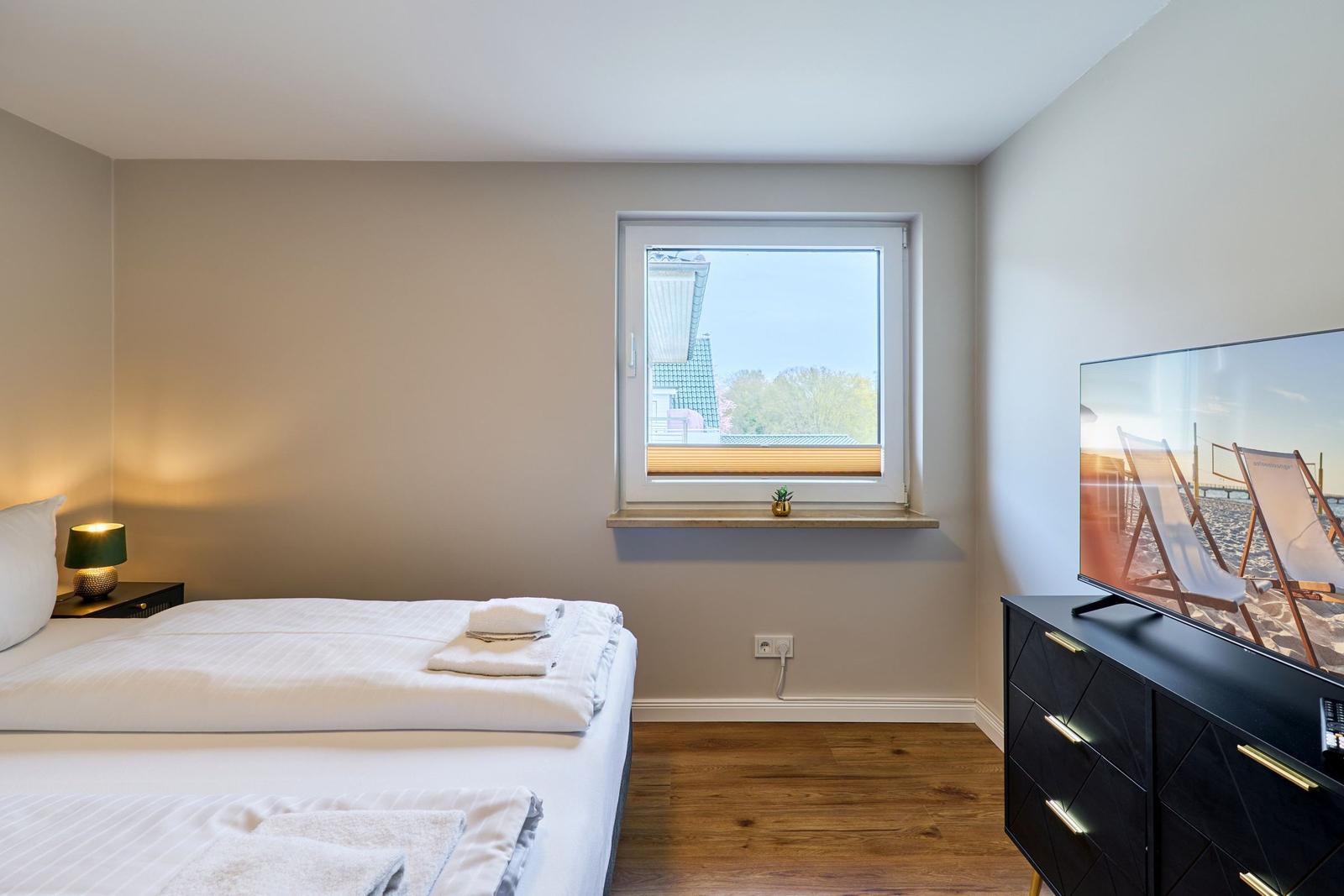 Schlafzimmer mit Bett, Fenster und Fernseher auf schwarzer Kommode.