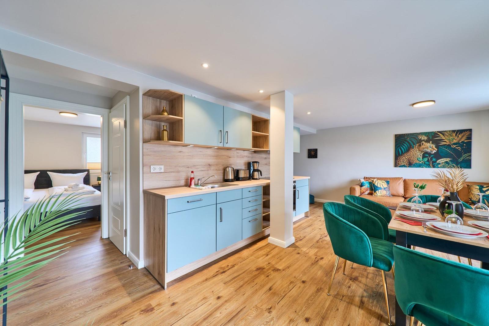Küche mit Essbereich und Schlafzimmer im Hintergrund. Grüne Stühle und Wandbild mit Leopard.