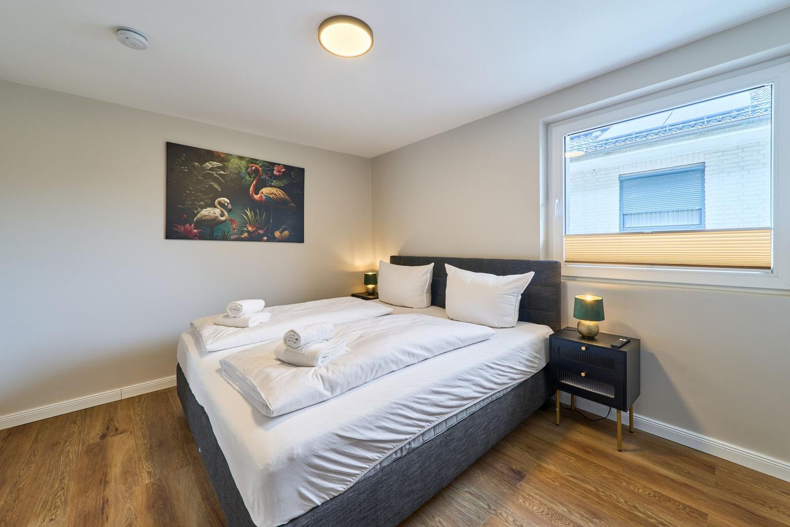 Schlafzimmer mit Bett, Nachttisch und Wandbild. Holzfußboden und Fenster mit Jalousie.