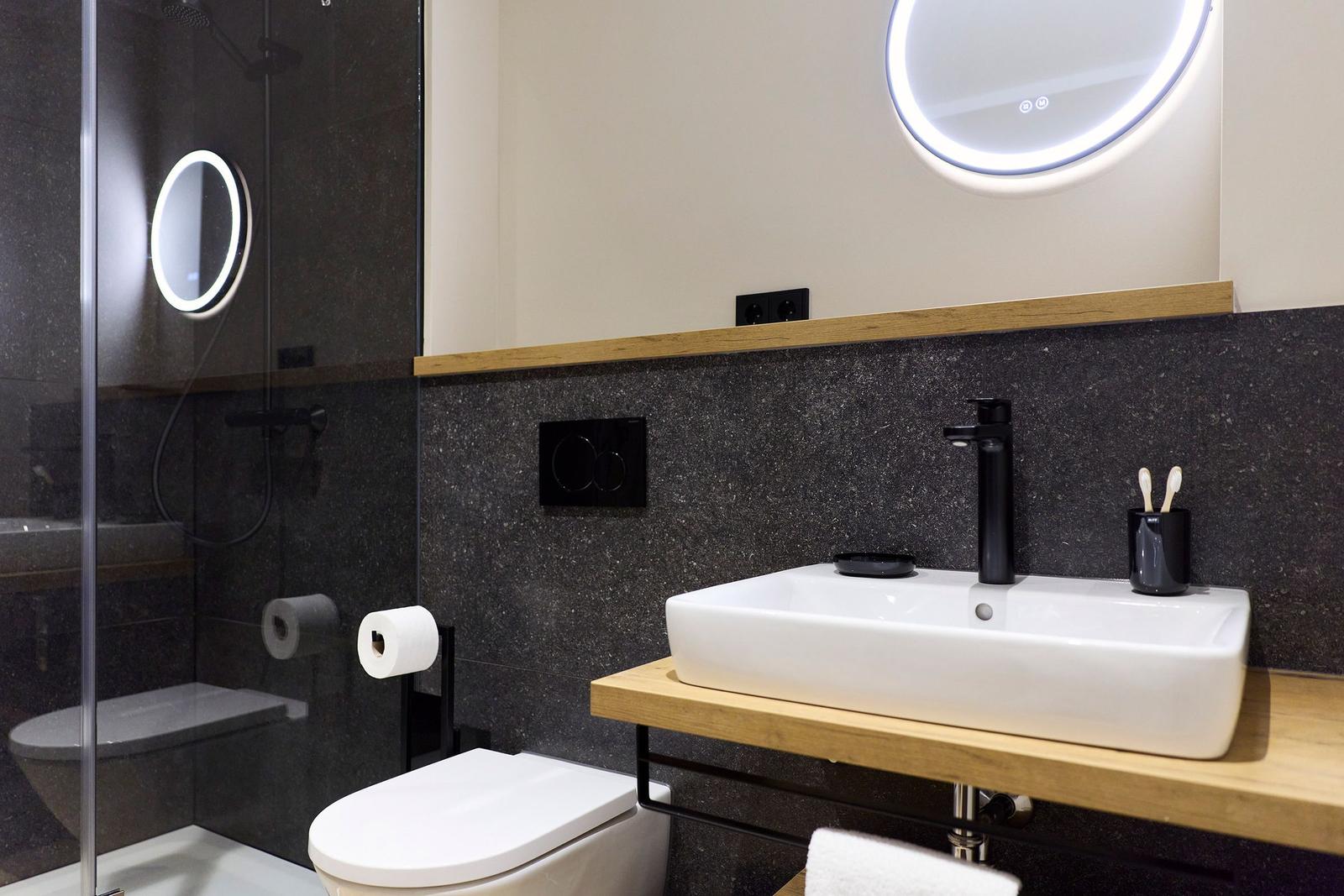 Badezimmer mit Dusche, Waschbecken und Toilette. Schwarze Wand und Holzoberflächen.