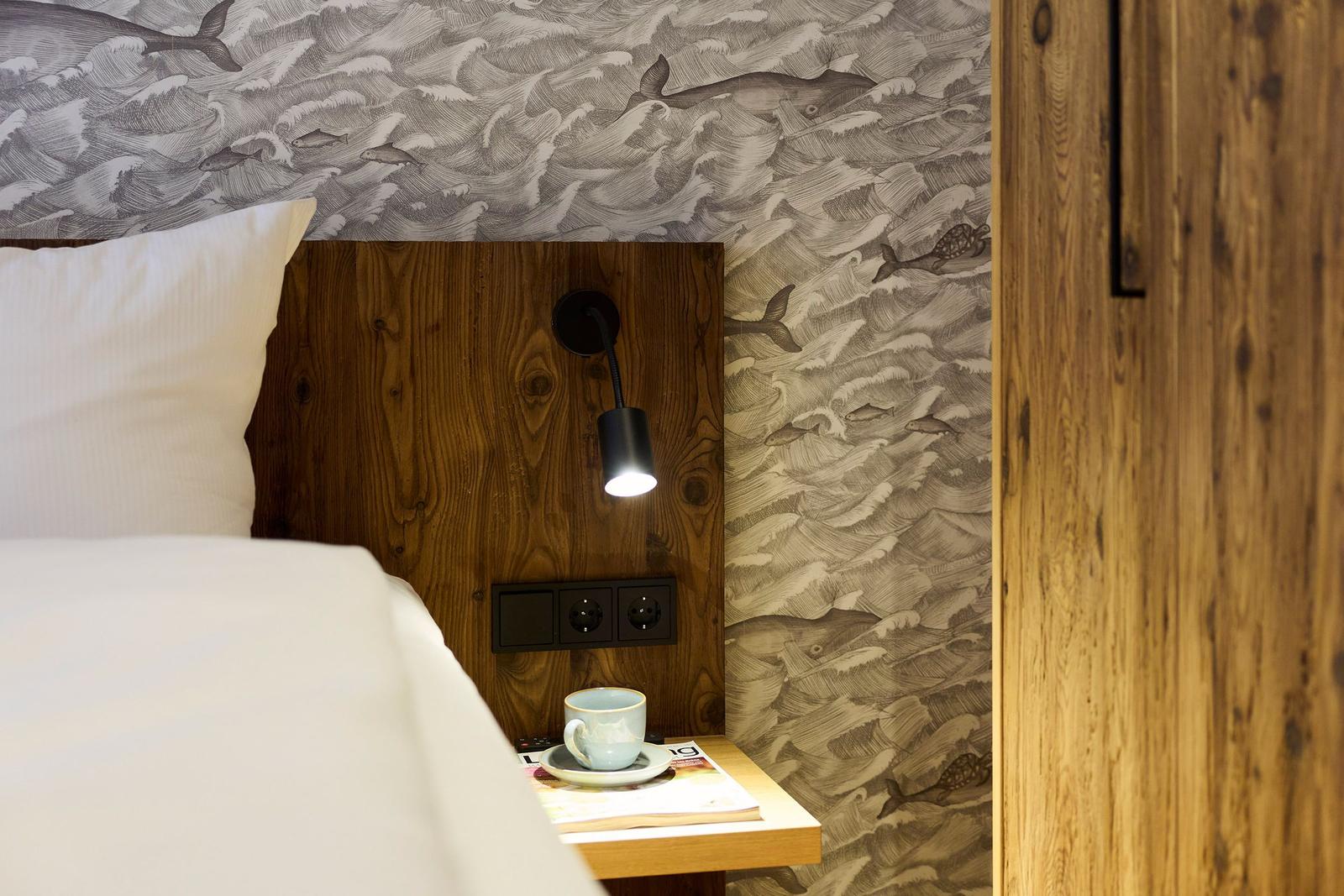 Schlafzimmer mit Holzkopfteil, Meerwasser-Wandtapete und Nachttisch mit Tasse.