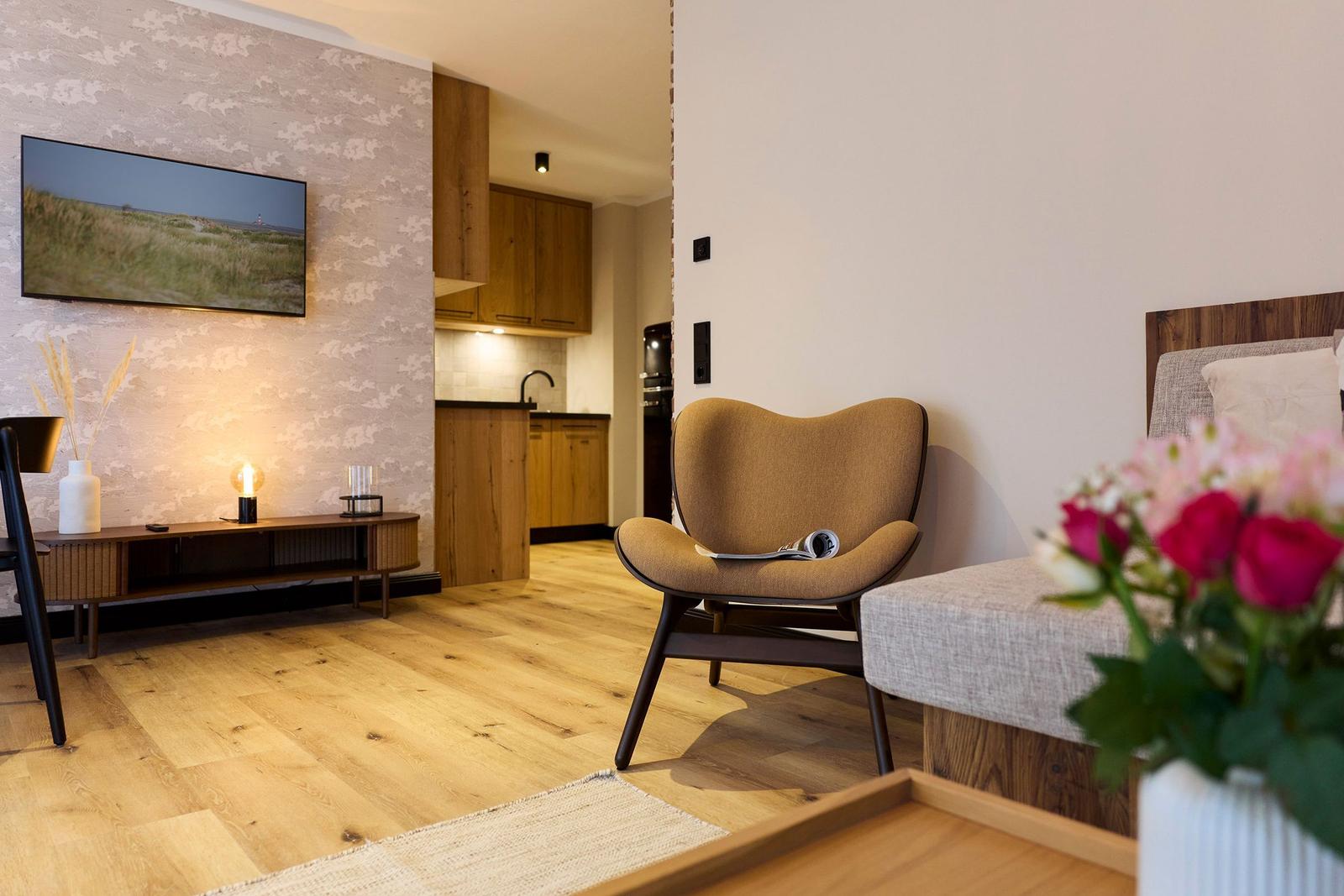 Wohnbereich mit Sessel, Fernseher und Küchenbereich. Holzboden und Blumen im Vordergrund.