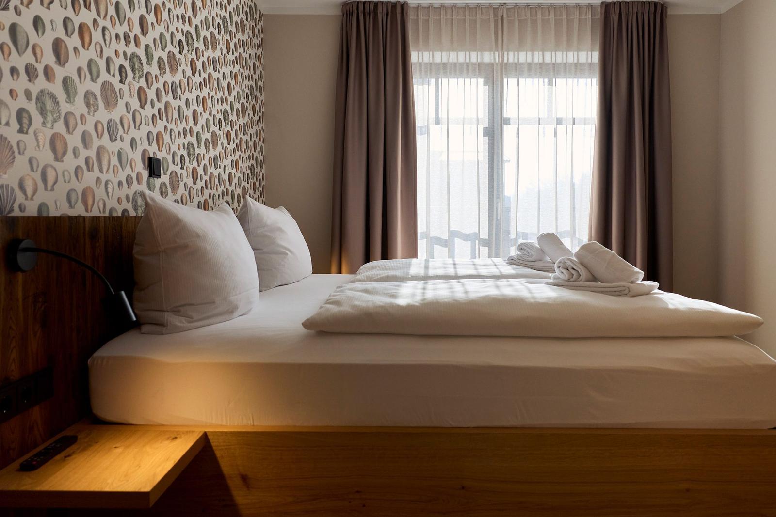 Ein Bett mit weißen Laken und Kissen, Wand mit Muschel-Muster und Fenster mit Vorhängen.