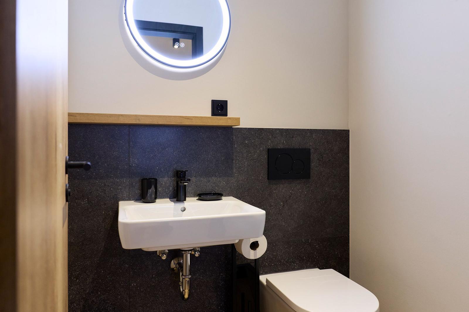 Badezimmer mit Waschbecken, Toilette und runden Spiegel mit Beleuchtung.