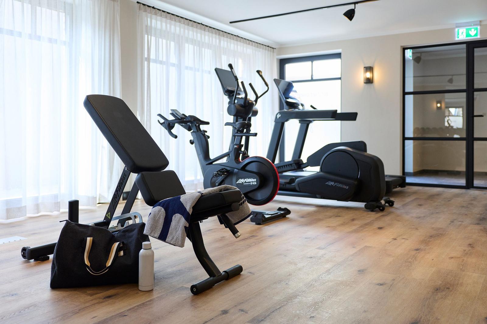 Fitnessraum mit Laufband, Crosstrainer und Bank. Holzboden und große Fenster.