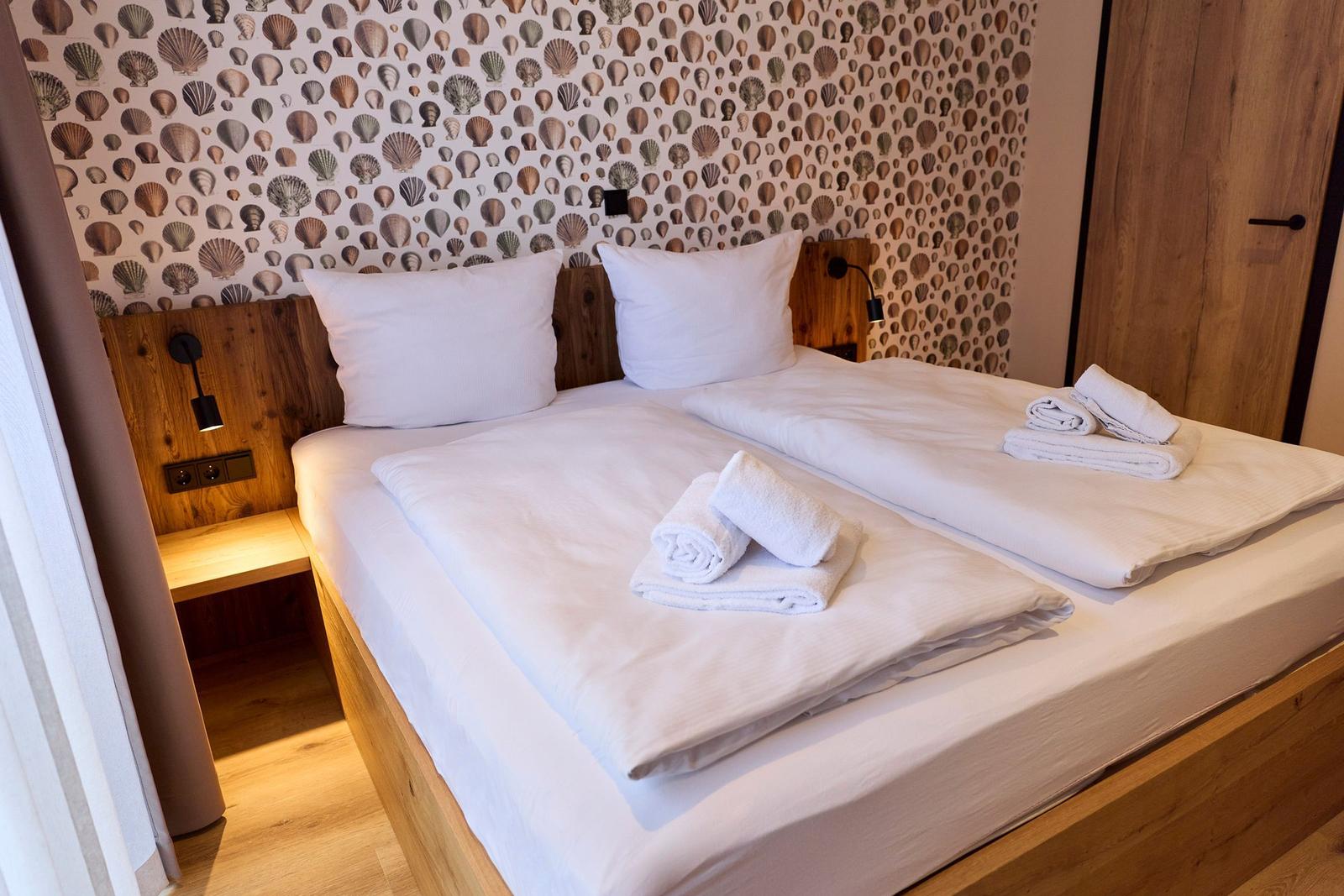 Doppelbett mit weißen Laken und Handtüchern, Wand mit Muschel-Motiv und Holzkopfteil.