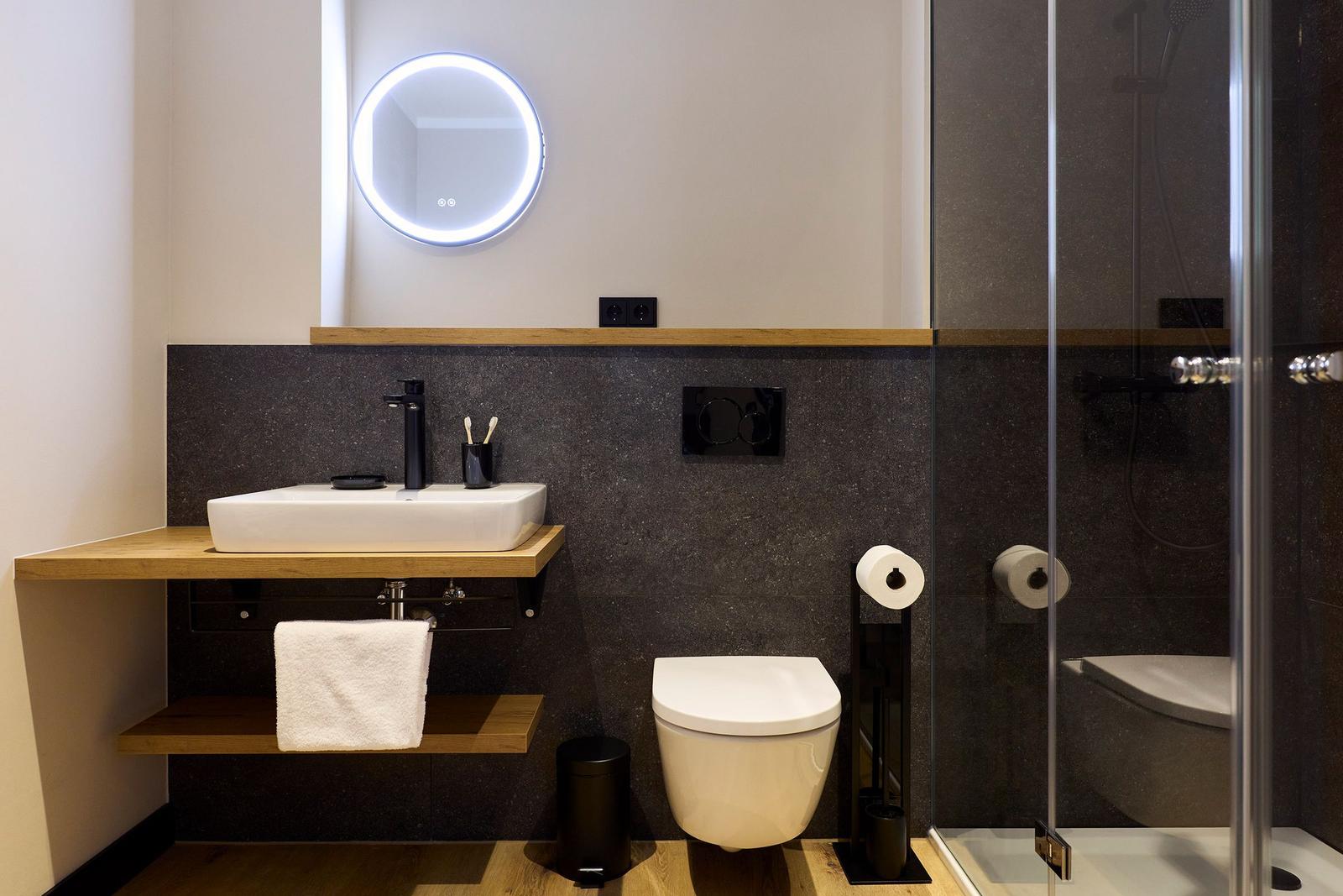 Badezimmer mit Waschtisch, Toilette und Dusche. Dunkle Wand, Holzregale und runder Spiegel.