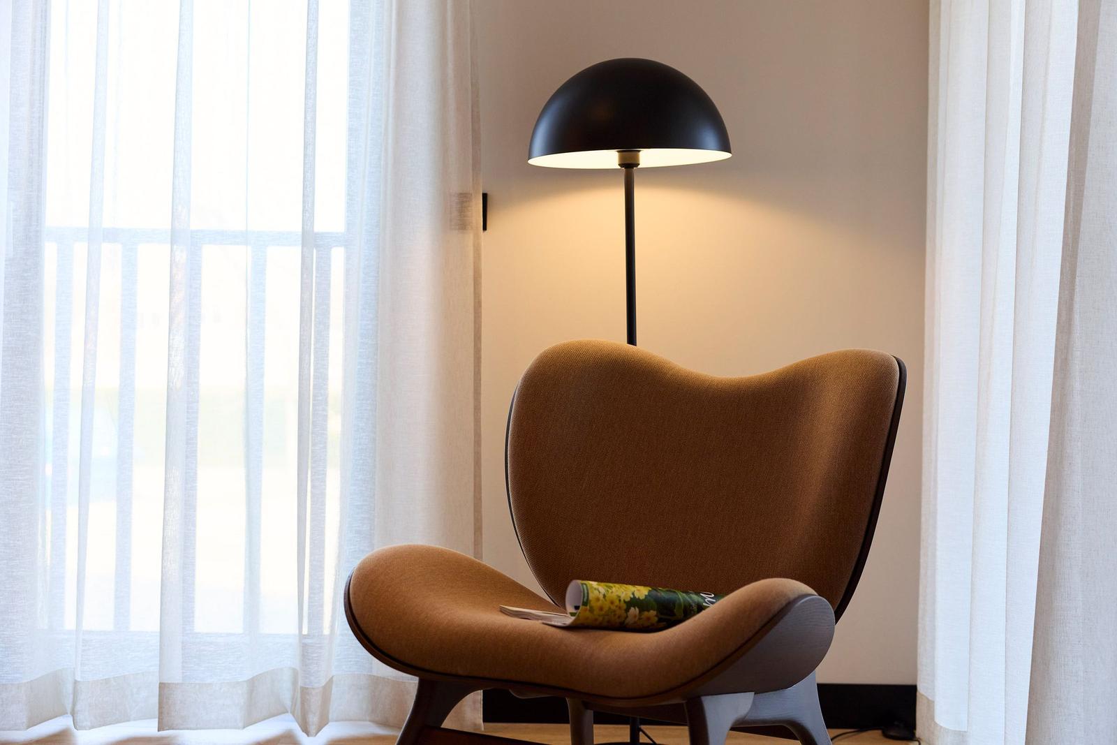 Ein braunes Sessel mit einem Buch und einer Stehlampe neben einem Fenster mit Vorhängen.