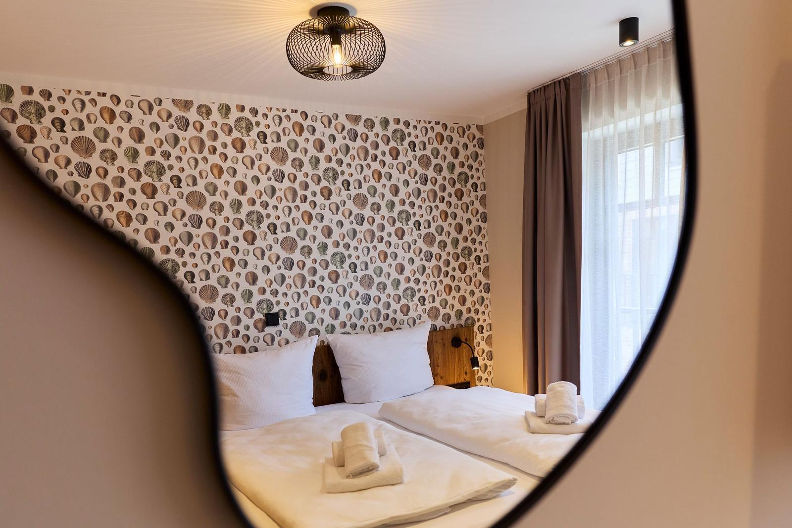 Zwei Betten mit weißen Laken und Handtüchern. Hintergrundwand mit Muschel-Motiv. Fenster mit Vorhängen.