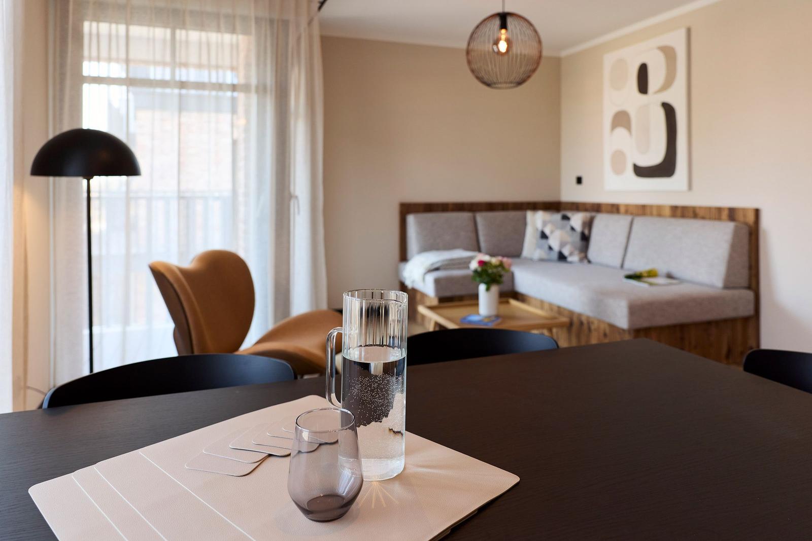 Esstisch mit Wasserglas und Karaffe im Essbereich. Im Hintergrund Sofa und Fenster.