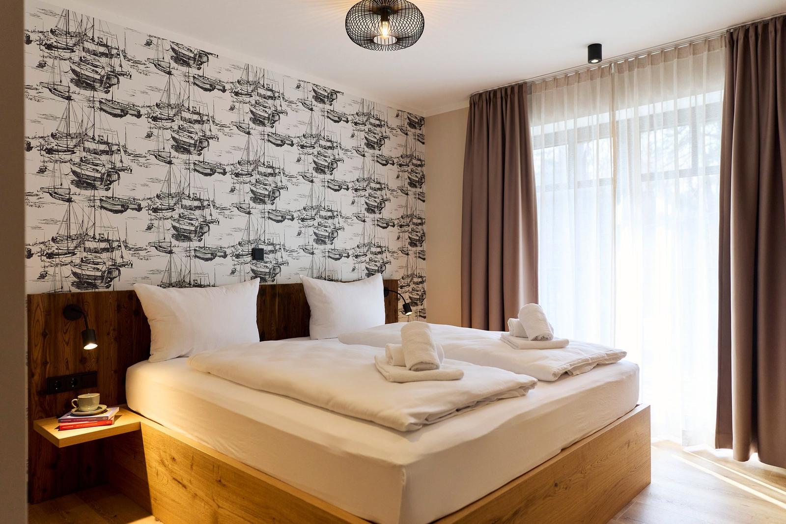 Schlafzimmer mit Bett, Holzgestell und Wandtapete mit Schiffsmotiven. Fenster mit Vorhängen.