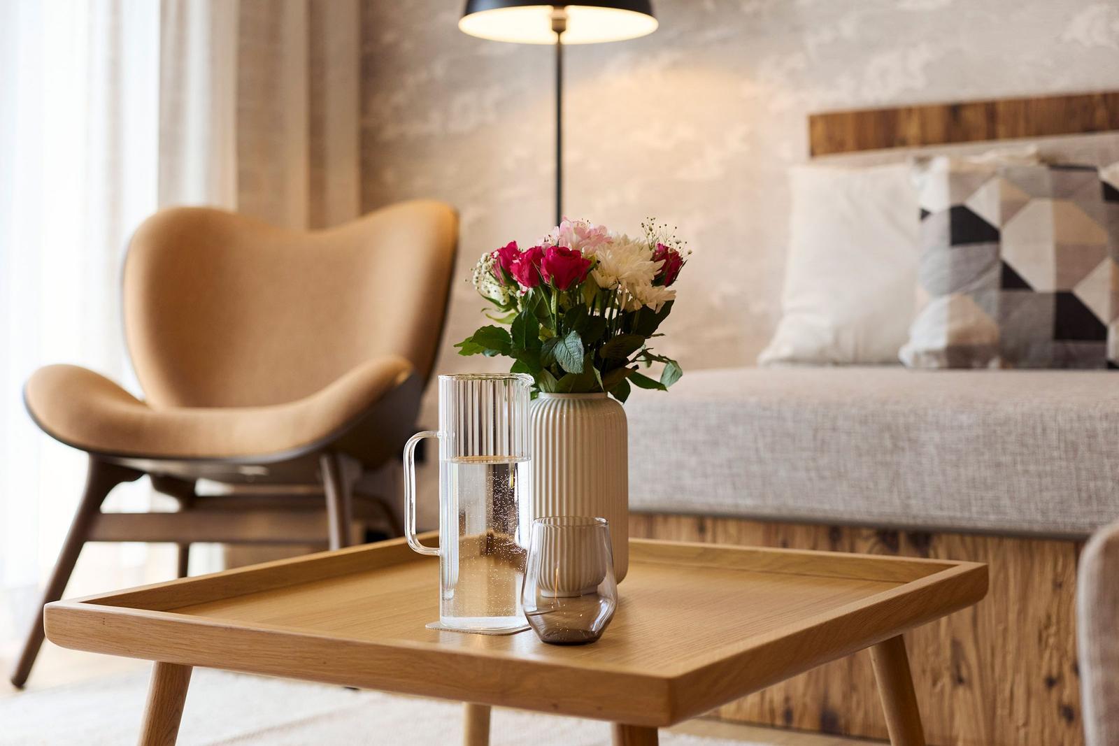 Ein Tisch mit Blumen, Wasserglas und Kerze vor einem Bett und Stuhl.