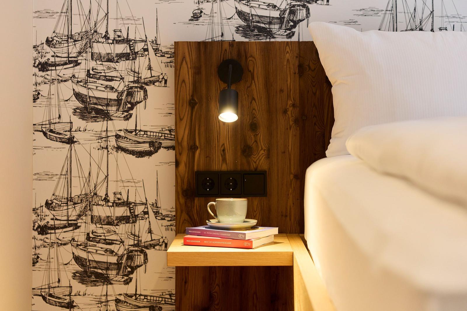 Schlafzimmer mit Holzkopfteil, Schiffsmotiv-Wandtapete und Nachttisch mit Tasse und Büchern.