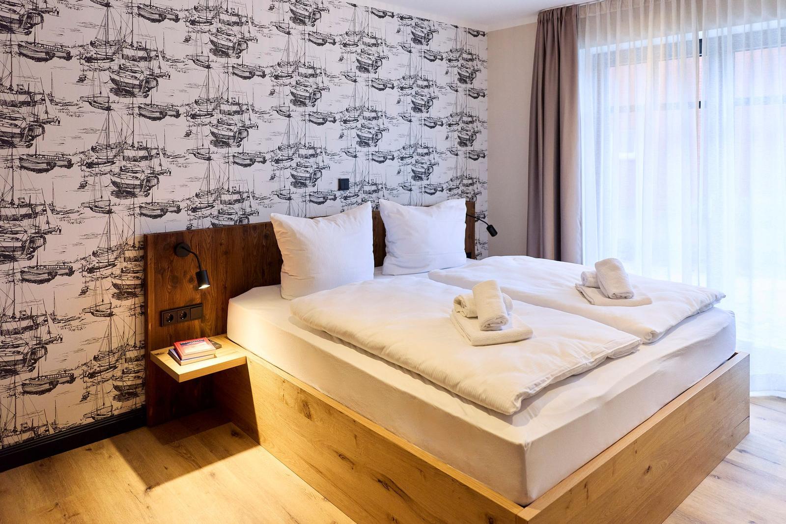Schlafzimmer mit Bett, Holzgestell und Segelyacht-Wandtapete. Fenster mit Vorhängen.