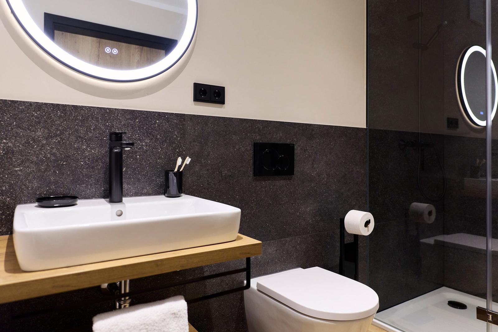 Bad mit Waschbecken, Toilette und Dusche. Spiegel mit Beleuchtung und Holzunterschrank.