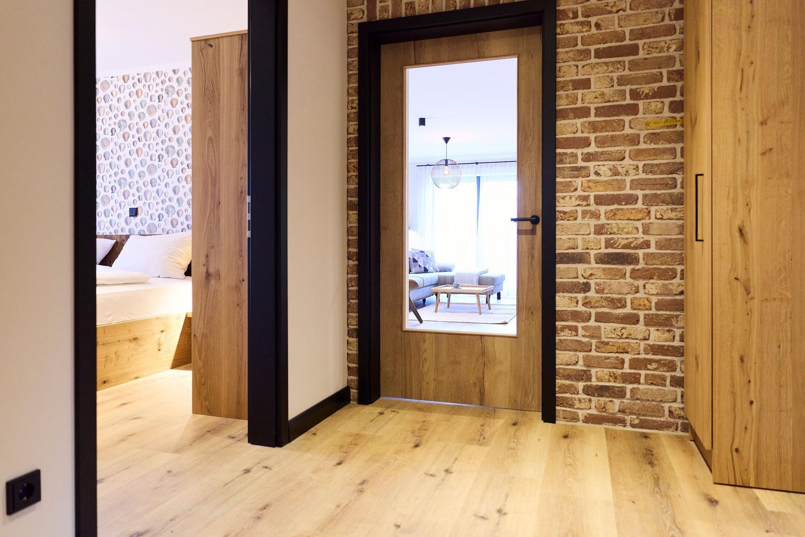 Schlafzimmer mit Muschelwand und Holzboden. Durch Tür ist Wohnbereich mit Sofa und Tisch sichtbar.