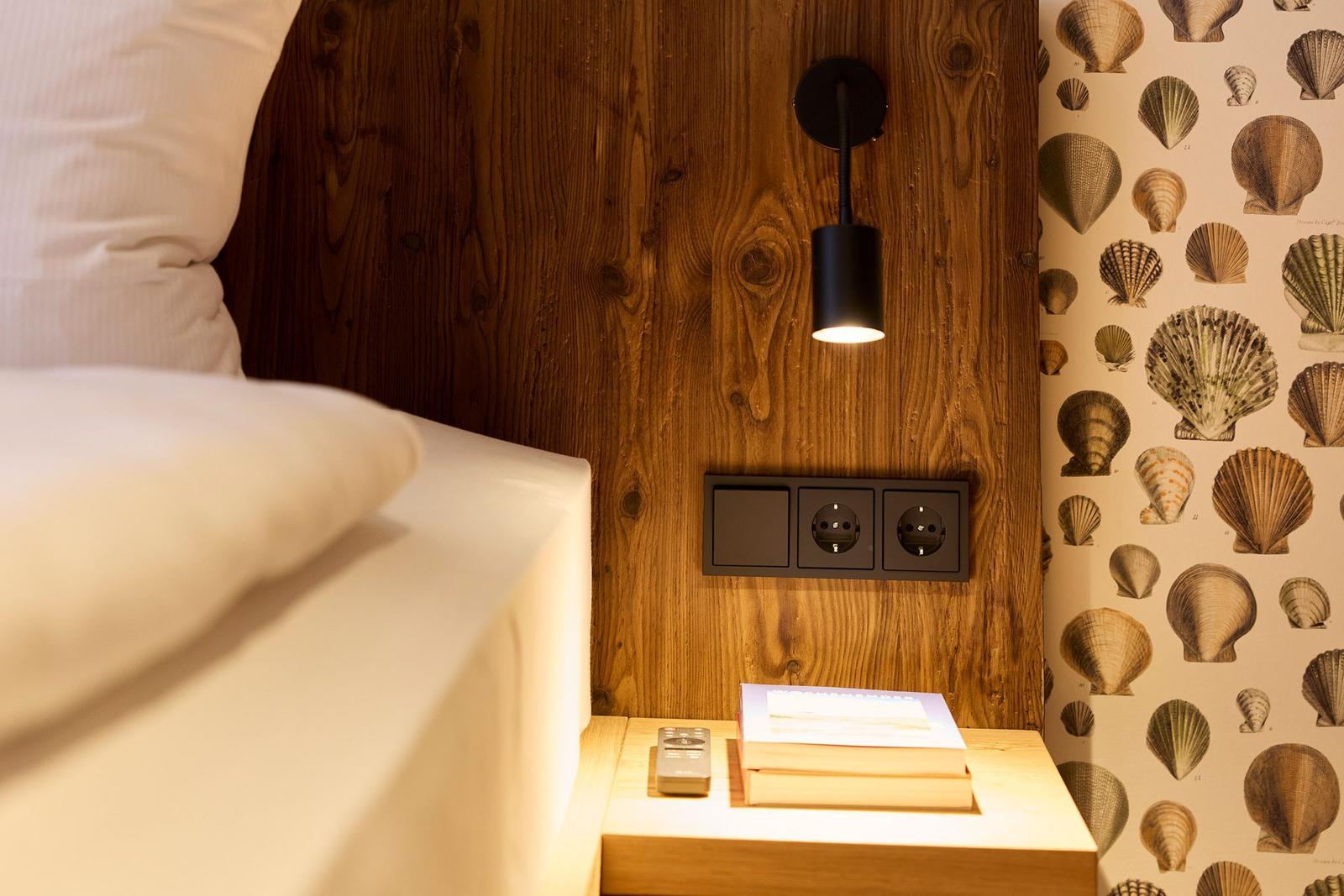 Bettkopfteil aus Holz mit Wandlampe und Steckdosen. Tapete mit Muschelmotiven. Nachttisch mit Buch und Fernbedienung.