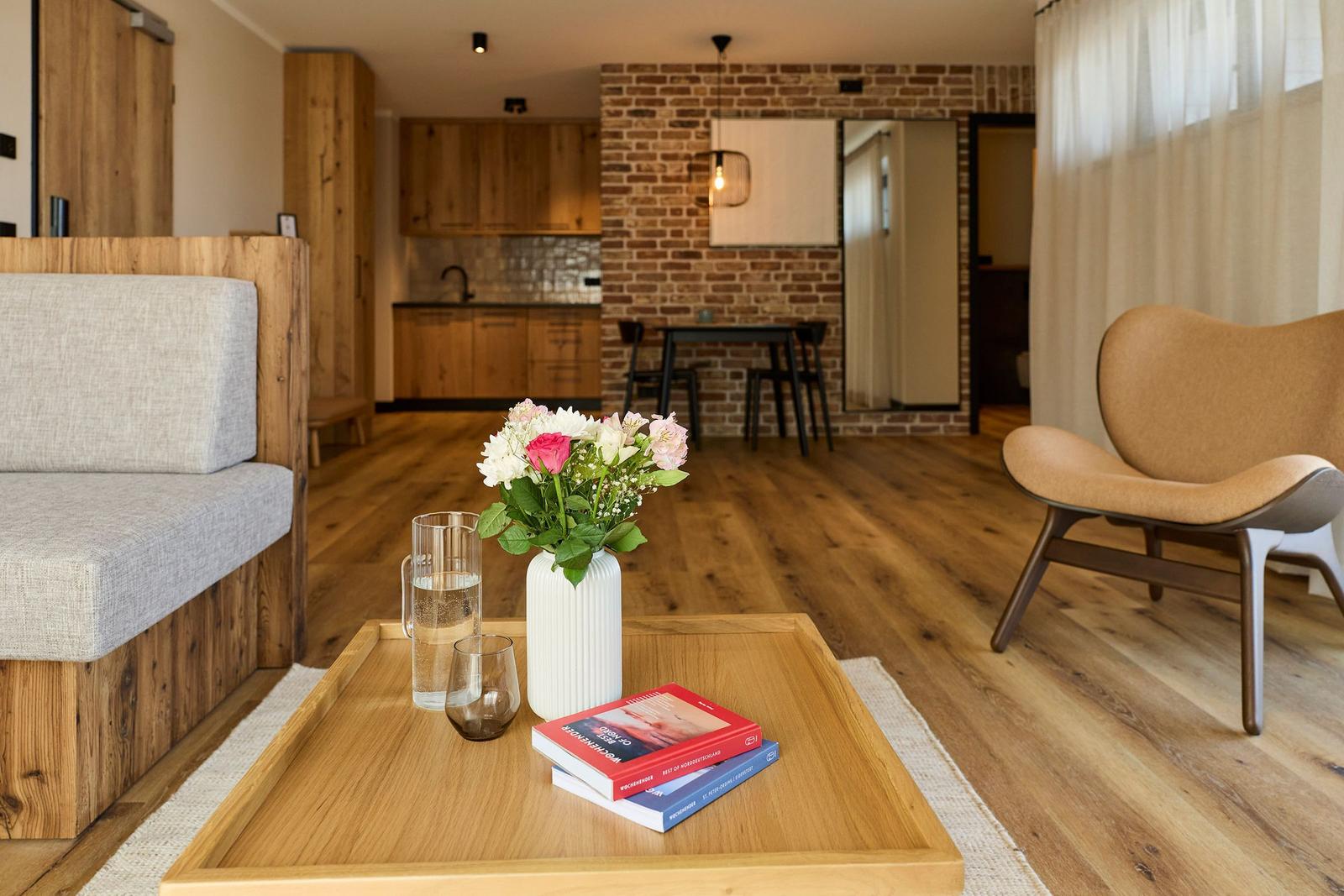 Wohnbereich mit Holzboden, Sofa, Stuhl und Tisch mit Blumen und Büchern.