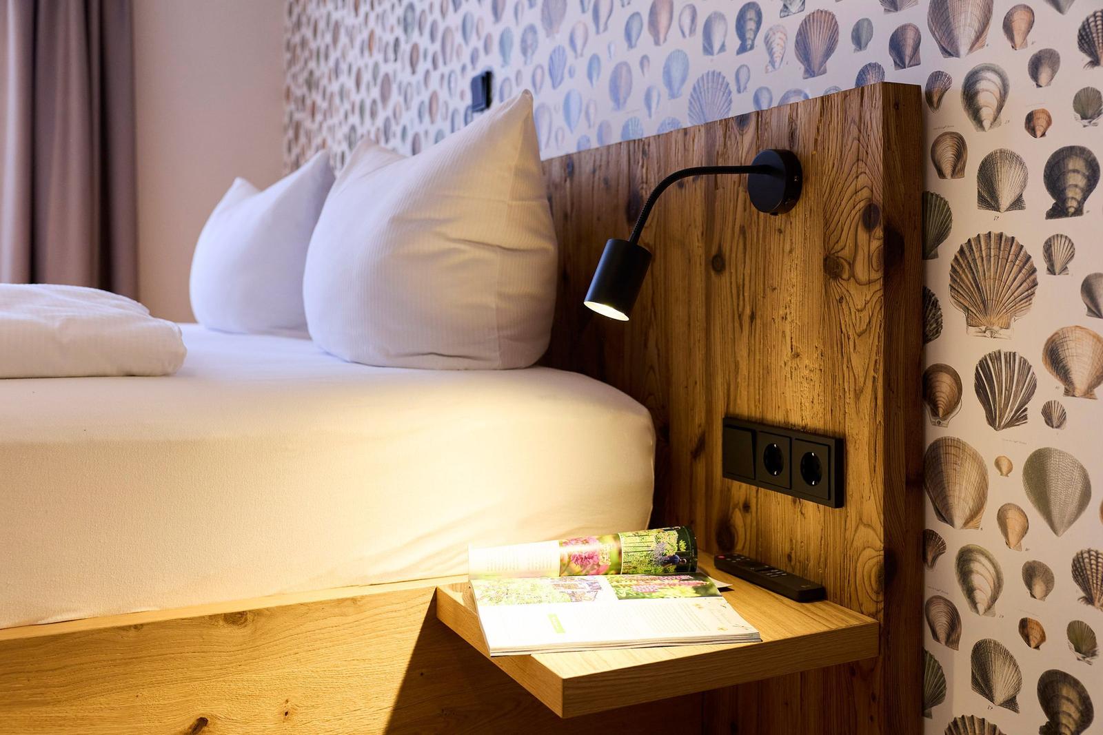 Bettkopf aus Holz mit Schaltflächen und Lampe. Wand mit Muschel-Motiv. Bett mit weißen Kissen.