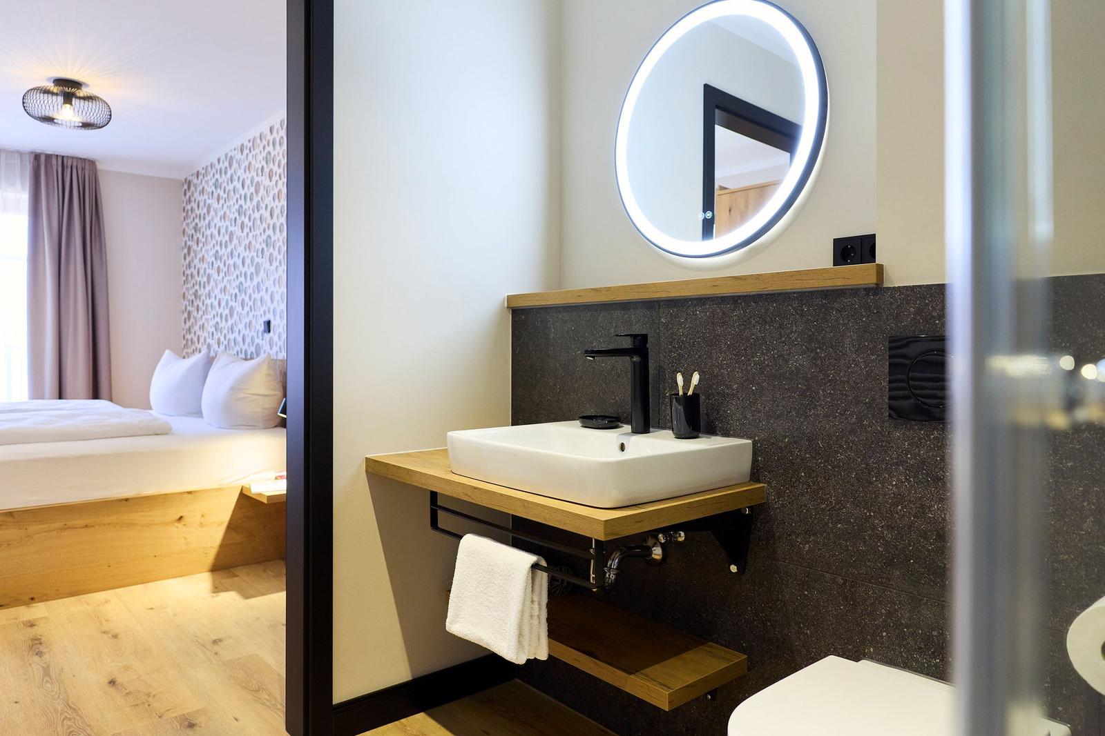 Badezimmer mit Waschbecken, Spiegel und Toilette. Im Hintergrund ist das Schlafzimmer zu sehen.