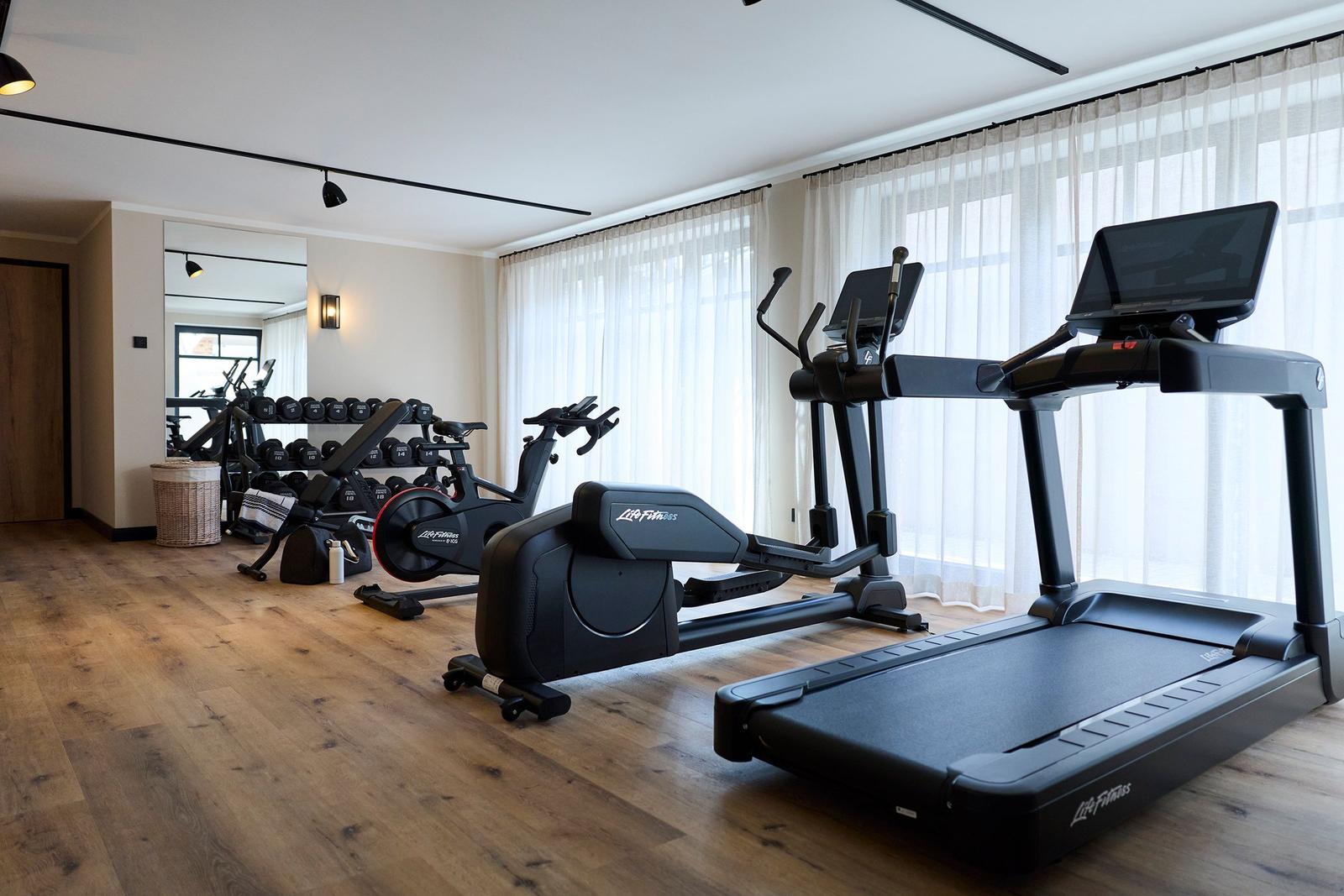 Fitnessraum mit Laufband, Ellipsentrainer und Fahrradtrainer. Holzboden und große Fenster.