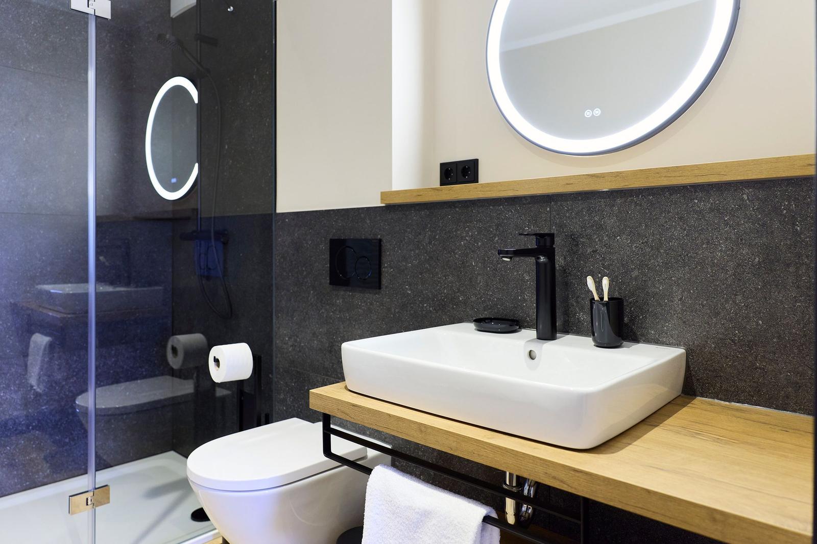 Badezimmer mit Dusche, Waschbecken und Toilette. Schwarze Wand, Holzoberfläche und beleuchtetes Spiegel.