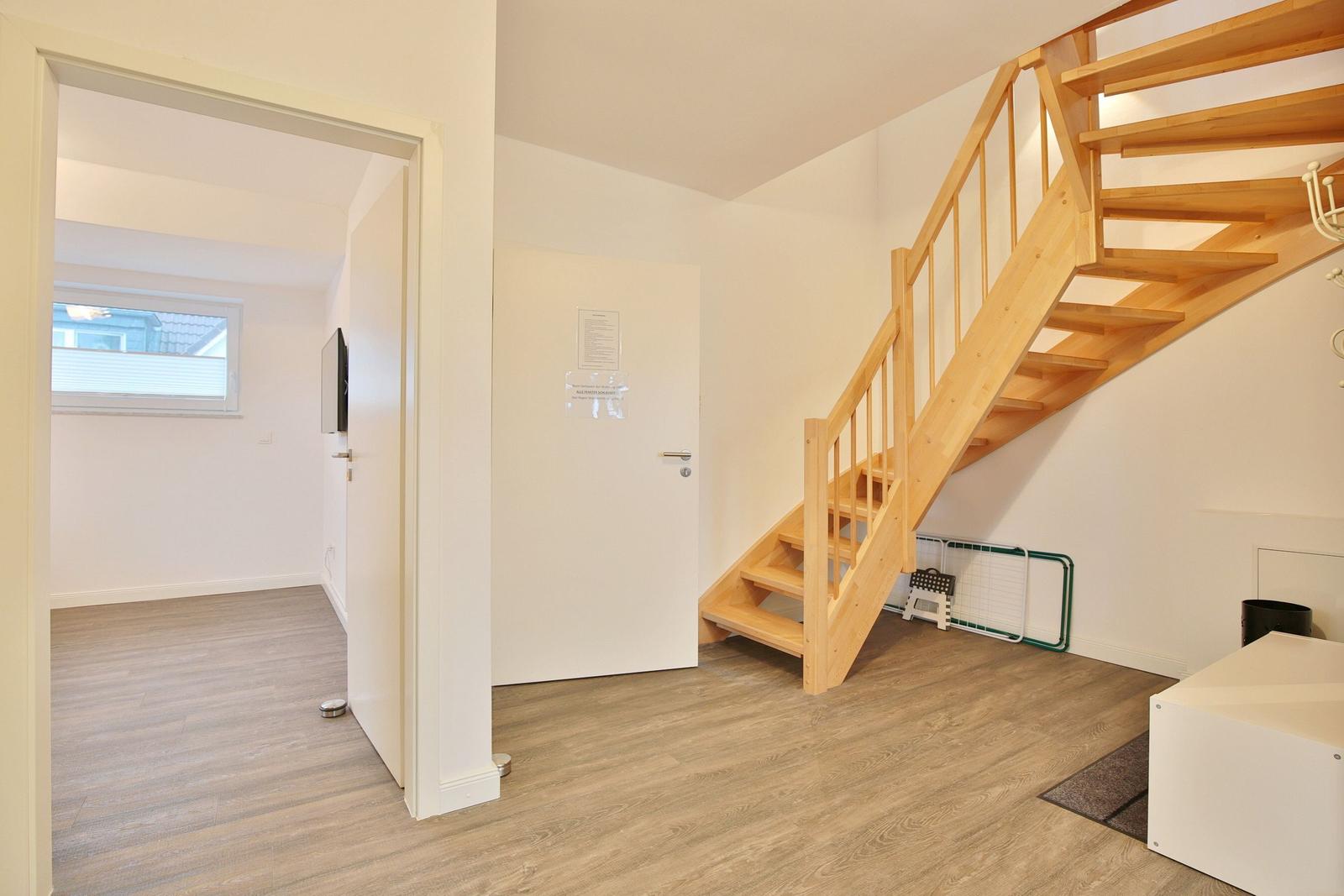 Eingangsbereich mit Holztreppe und Tür zu Wohnraum mit Fenster und Fernseher.