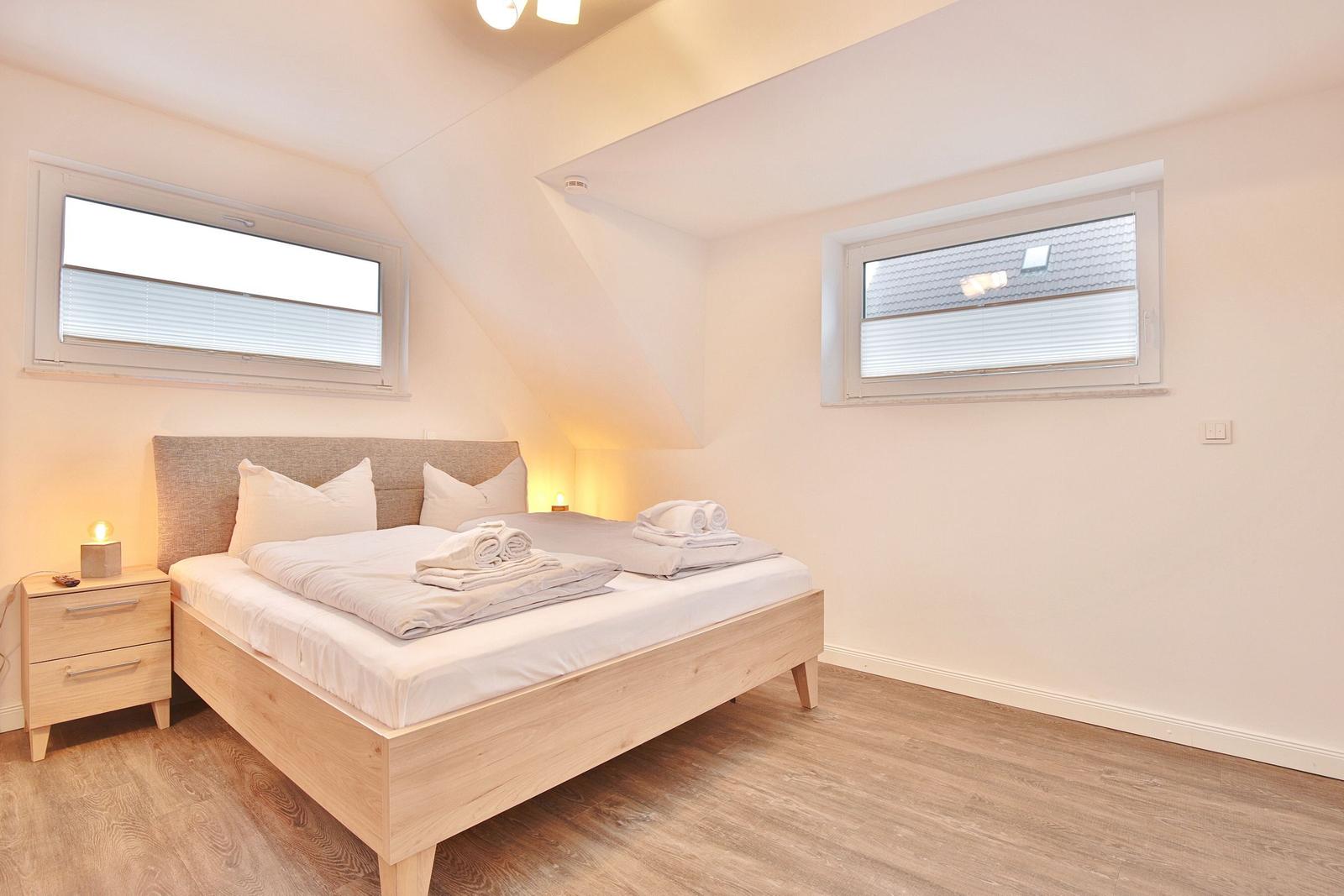 Schlafzimmer mit Doppelbett, zwei Fenstern und Holzboden.