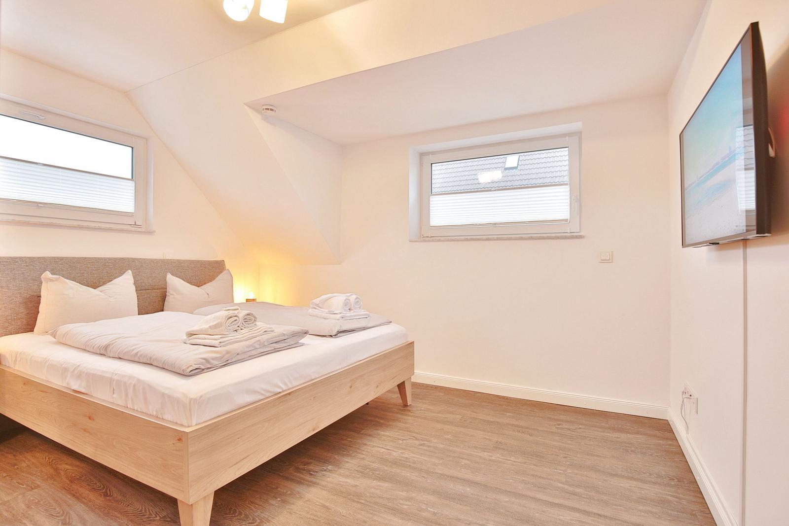 Schlafzimmer mit Doppelbett, zwei Fenstern und Fernseher an der Wand.