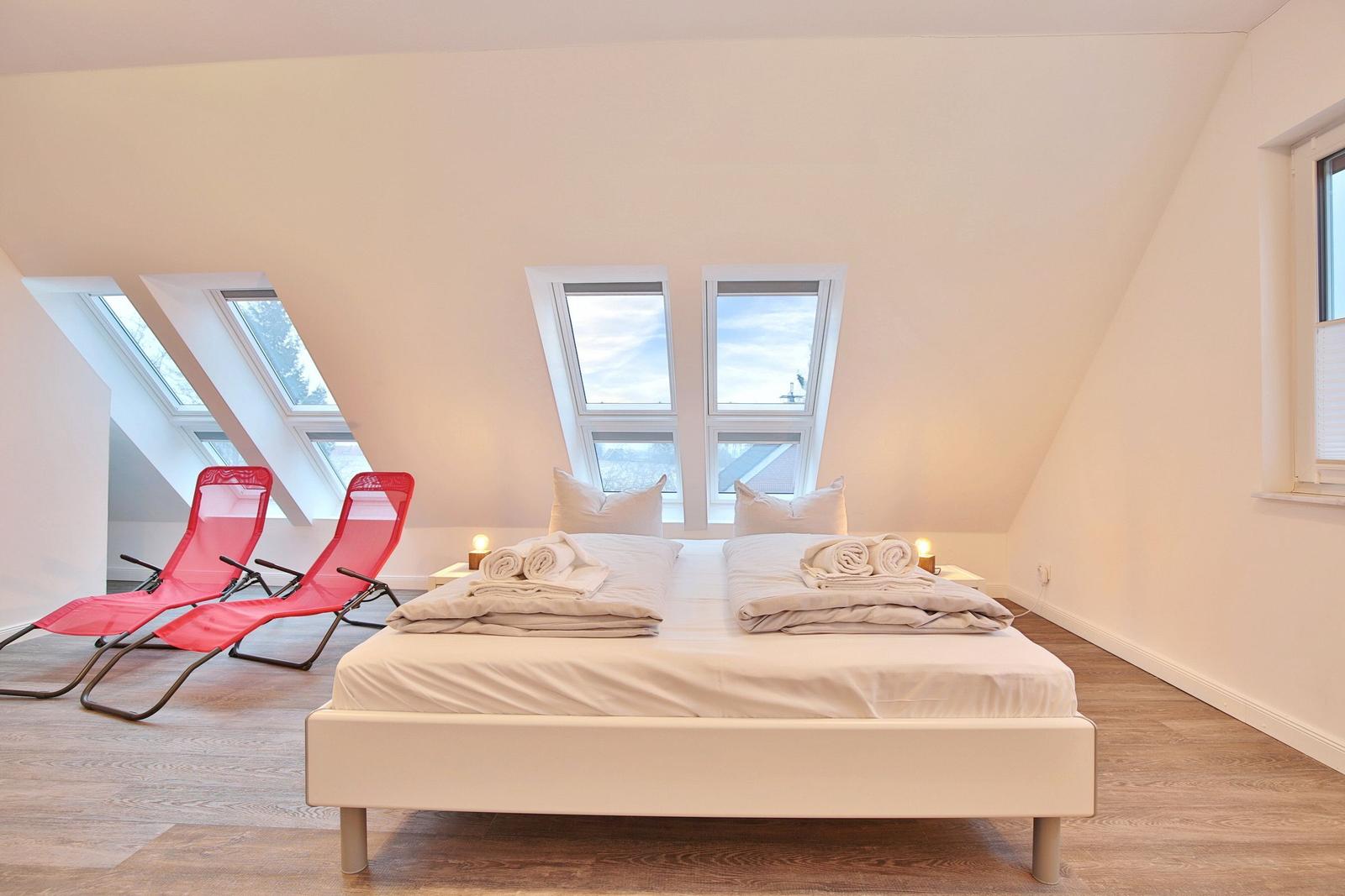 Schlafzimmer mit Dachfenstern, Bett und zwei roten Liegestühlen.