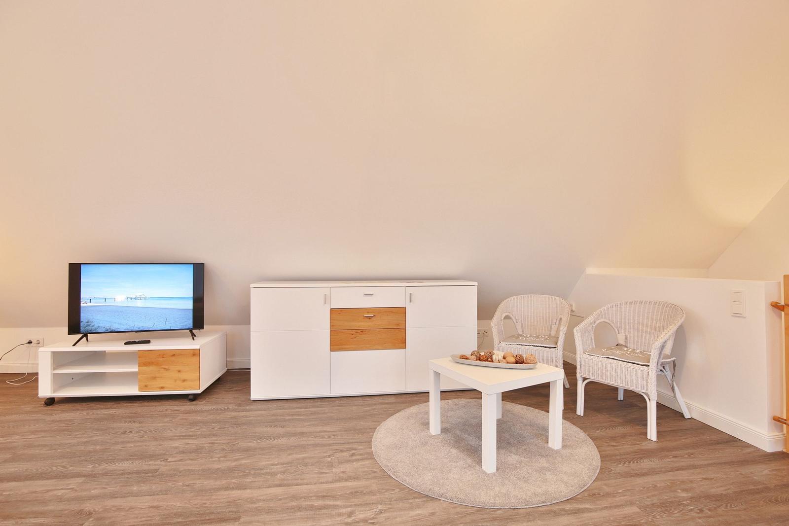 Wohnzimmer mit Fernseher, Sideboard, zwei Stühlen und Tisch auf Teppich.