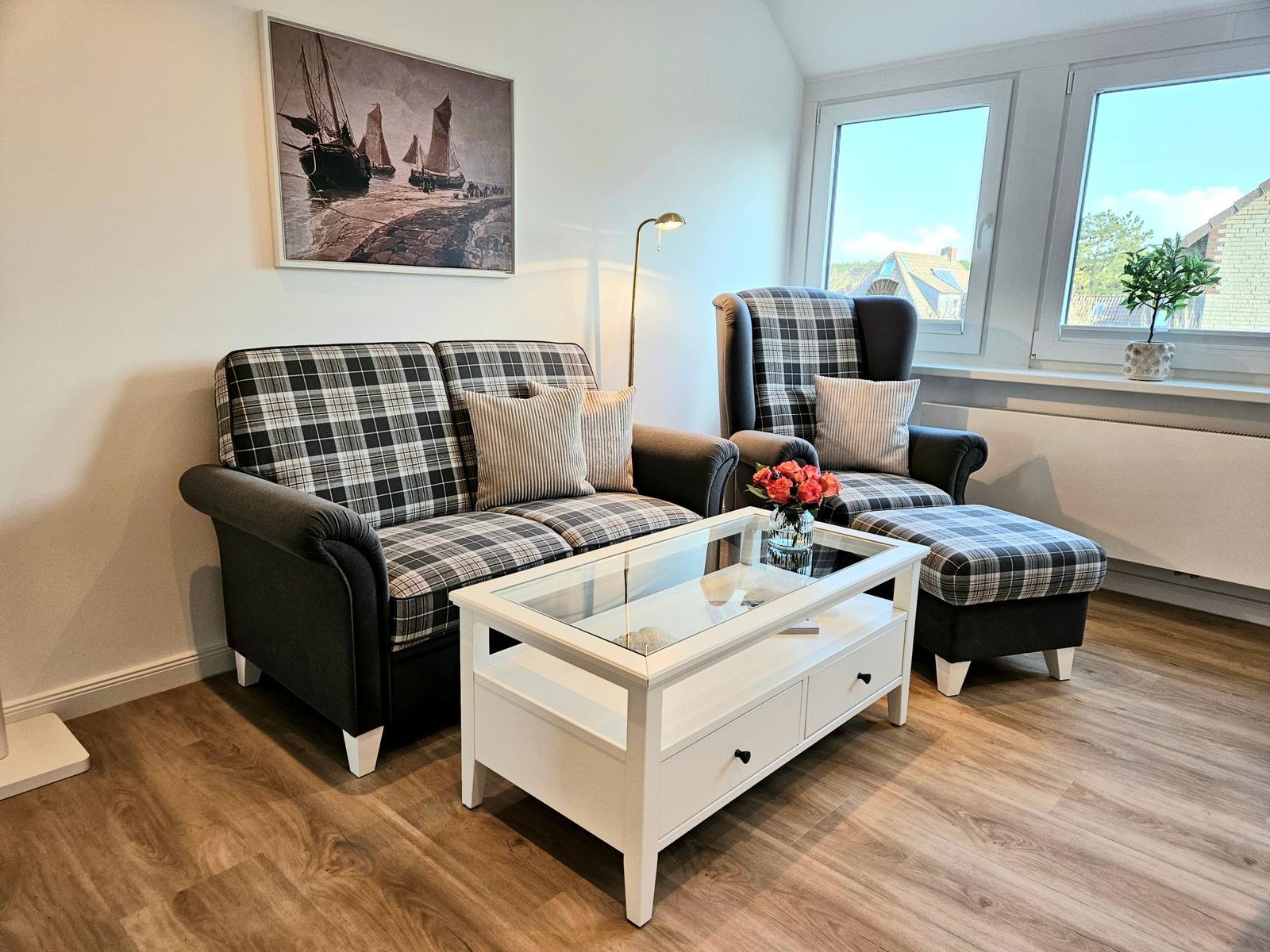 Wohnzimmer mit Sofa, Sessel und Couchtisch. An der Wand hängt ein Bild mit Schiffen.