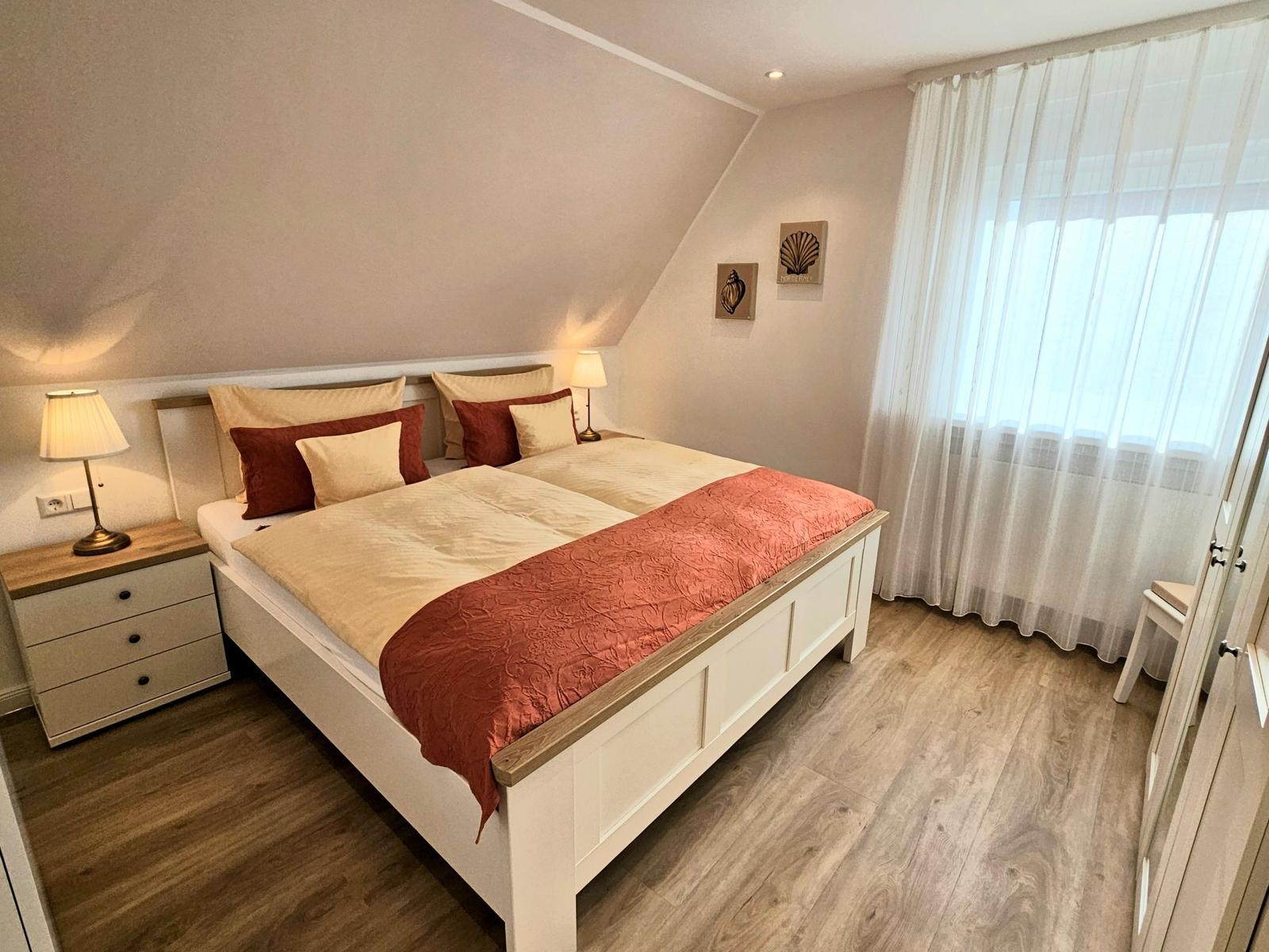 Schlafzimmer mit Doppelbett, Nachttisch, Lampe und Fenster mit Vorhängen.