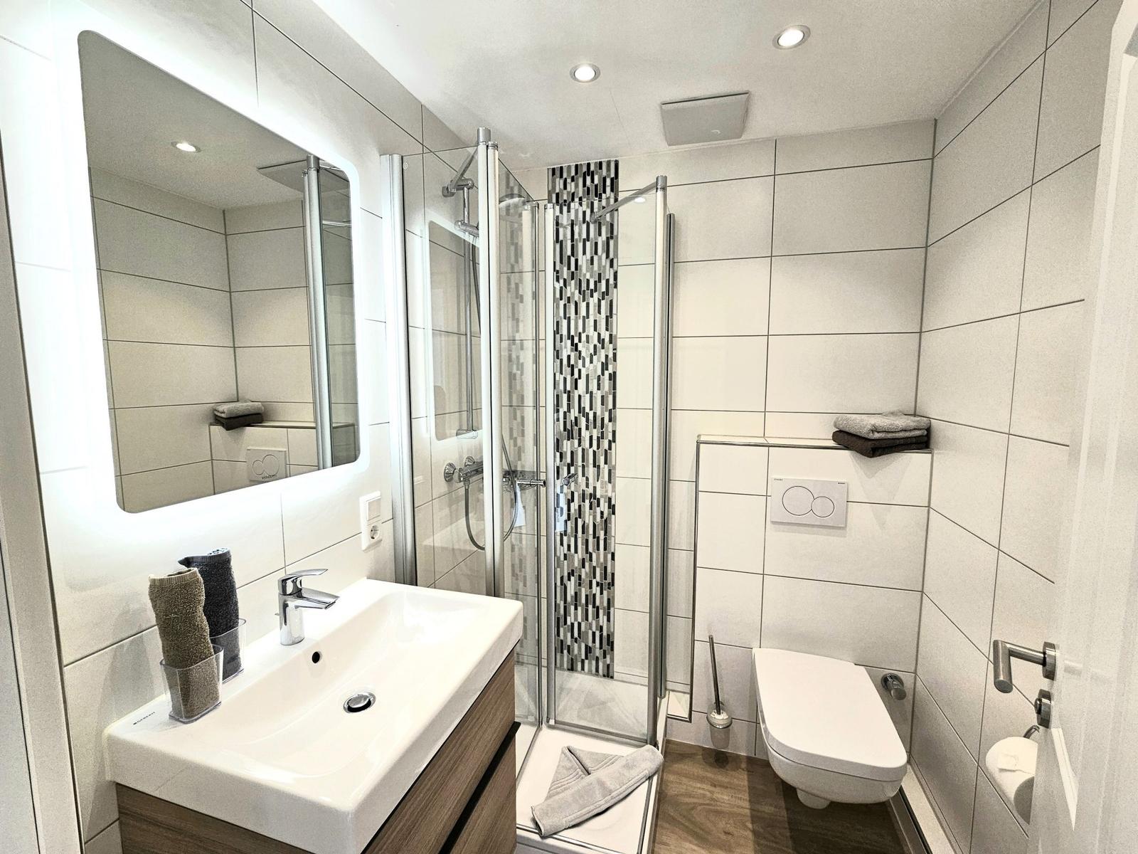 Badezimmer mit Dusche, Waschbecken und Toilette. Fliesenwand mit Mosaikverkleidung.