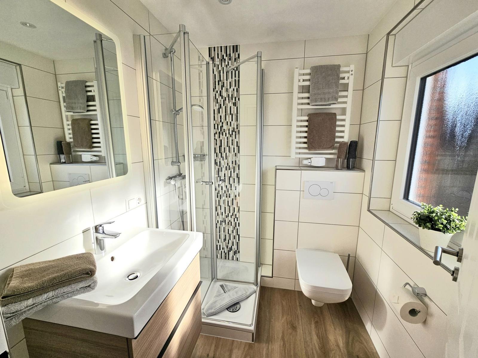 Badezimmer mit Dusche, Waschbecken und Toilette. Fenster mit Pflanze.