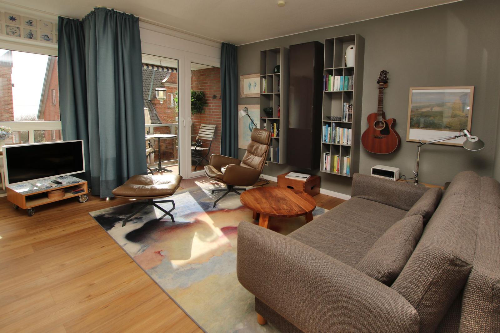 Wohnzimmer mit Sofa, Sessel, Bücherregal und Fernseher. Blick auf Balkon mit Tisch und Stühlen.