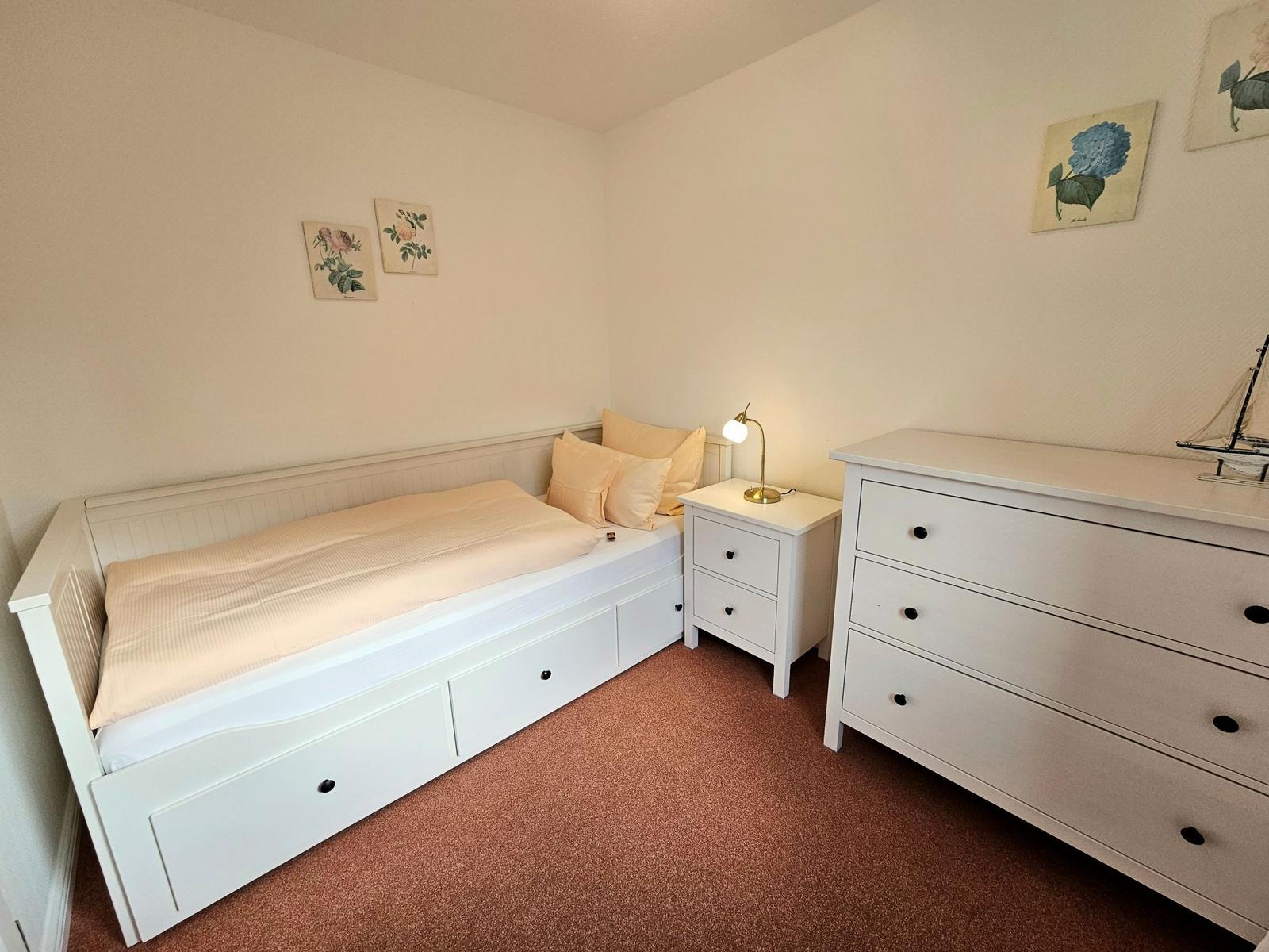 Bedroom with white bed, nightstand, and dresser. Wall art and carpeted floor.