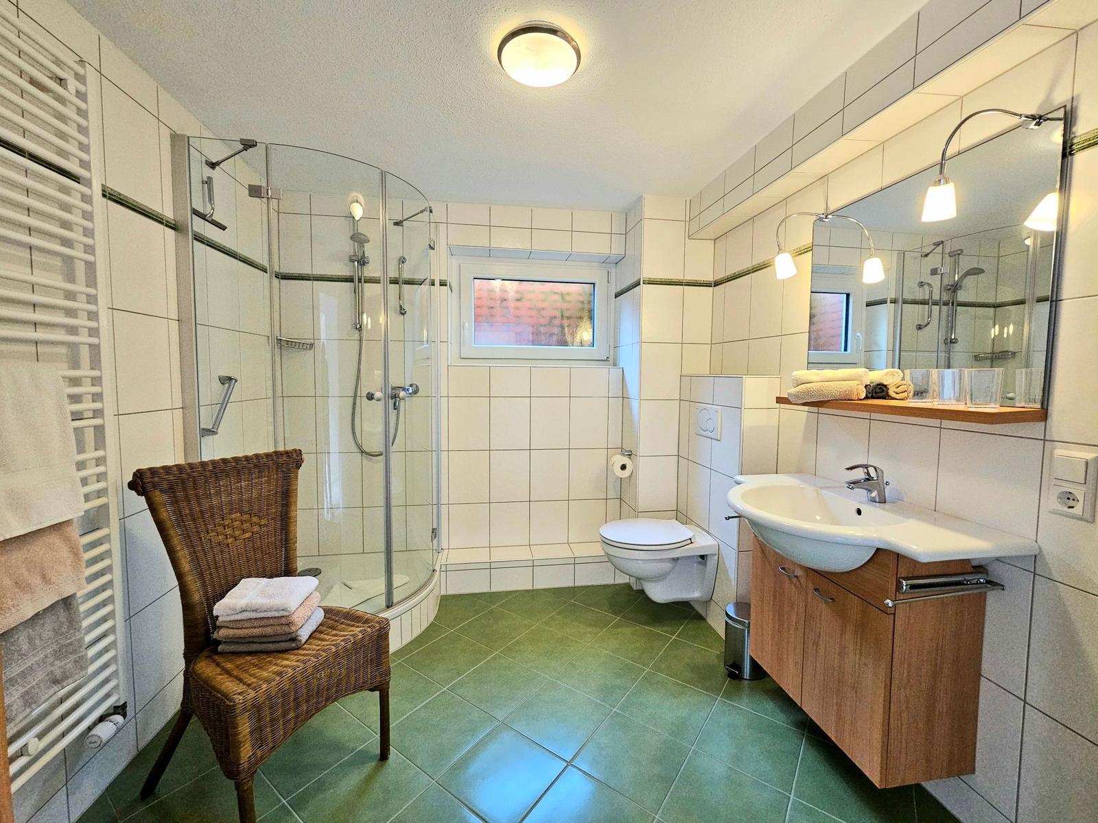 Bathroom with shower, sink, and toilet. Green tiled floor. Wicker chair with towels.