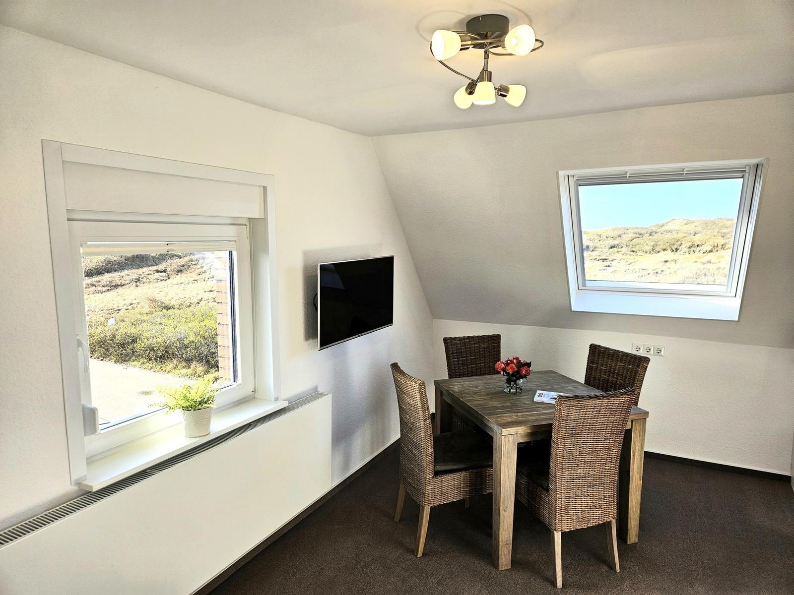 Esszimmer mit Tisch, vier Stühlen und TV. Zwei Fenster bieten Blick auf Landschaft.